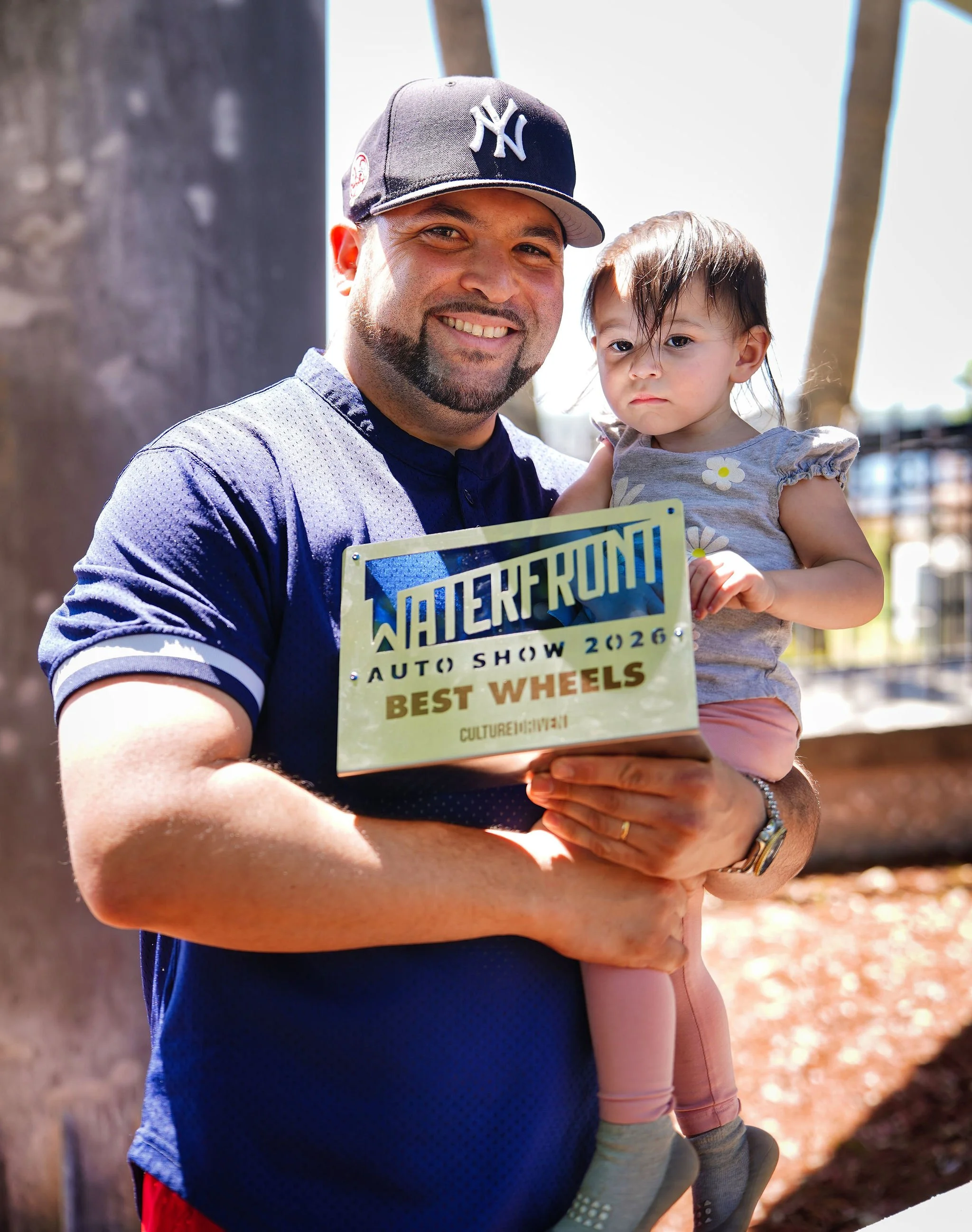 A man in a baseball cap holding a young girl, both smiling. The man is holding a plaque that reads 'Waterfront Auto Show 2026 Best Wheels.'