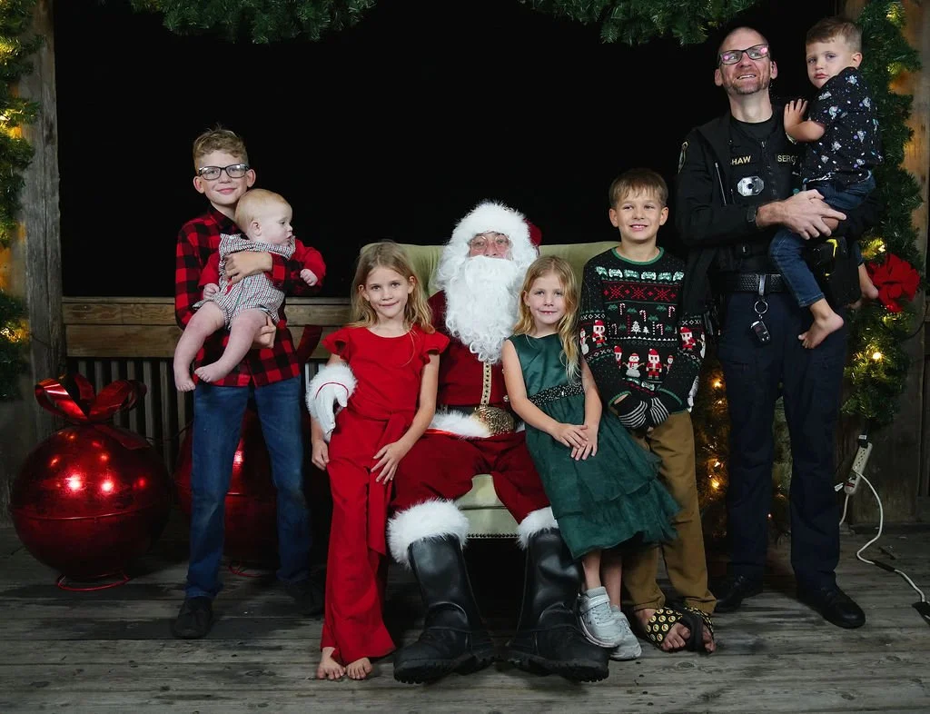 Children and adults with Santa Claus sitting and standing in front of Christmas decorations, including a large red ornament and a Christmas tree with lights and ornaments.