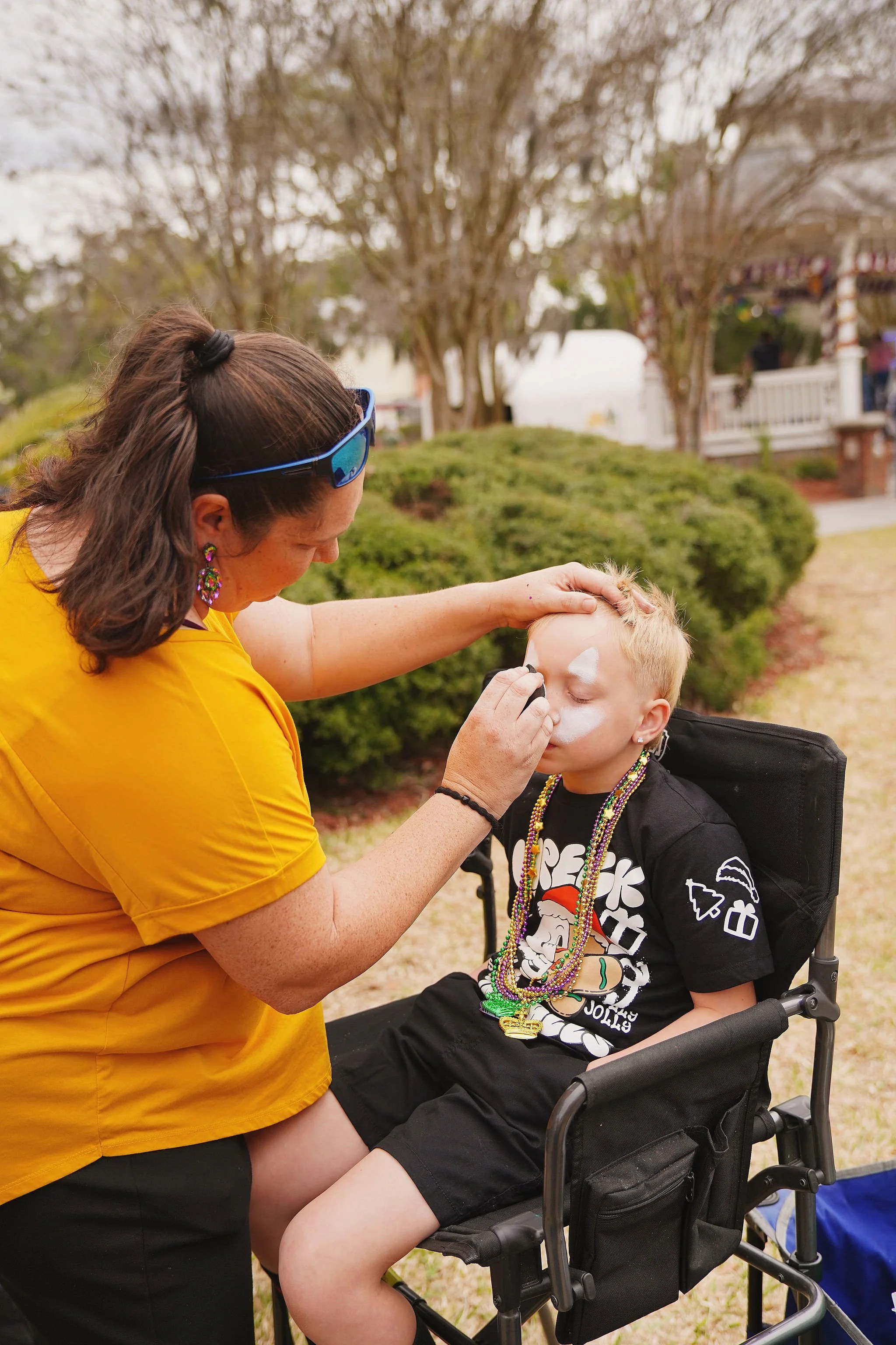 A woman in a yellow shirt is painting a boy's face white while he sits in a wheelchair. The boy is wearing a black T-shirt with a graphic design and has colorful beads around his neck.
