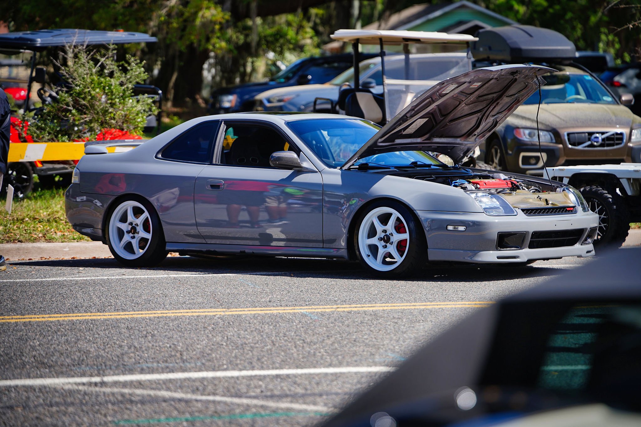 A silver Acura NSX with the hood open, parked in a lot during a car event. Several other cars and structures are visible in the background.