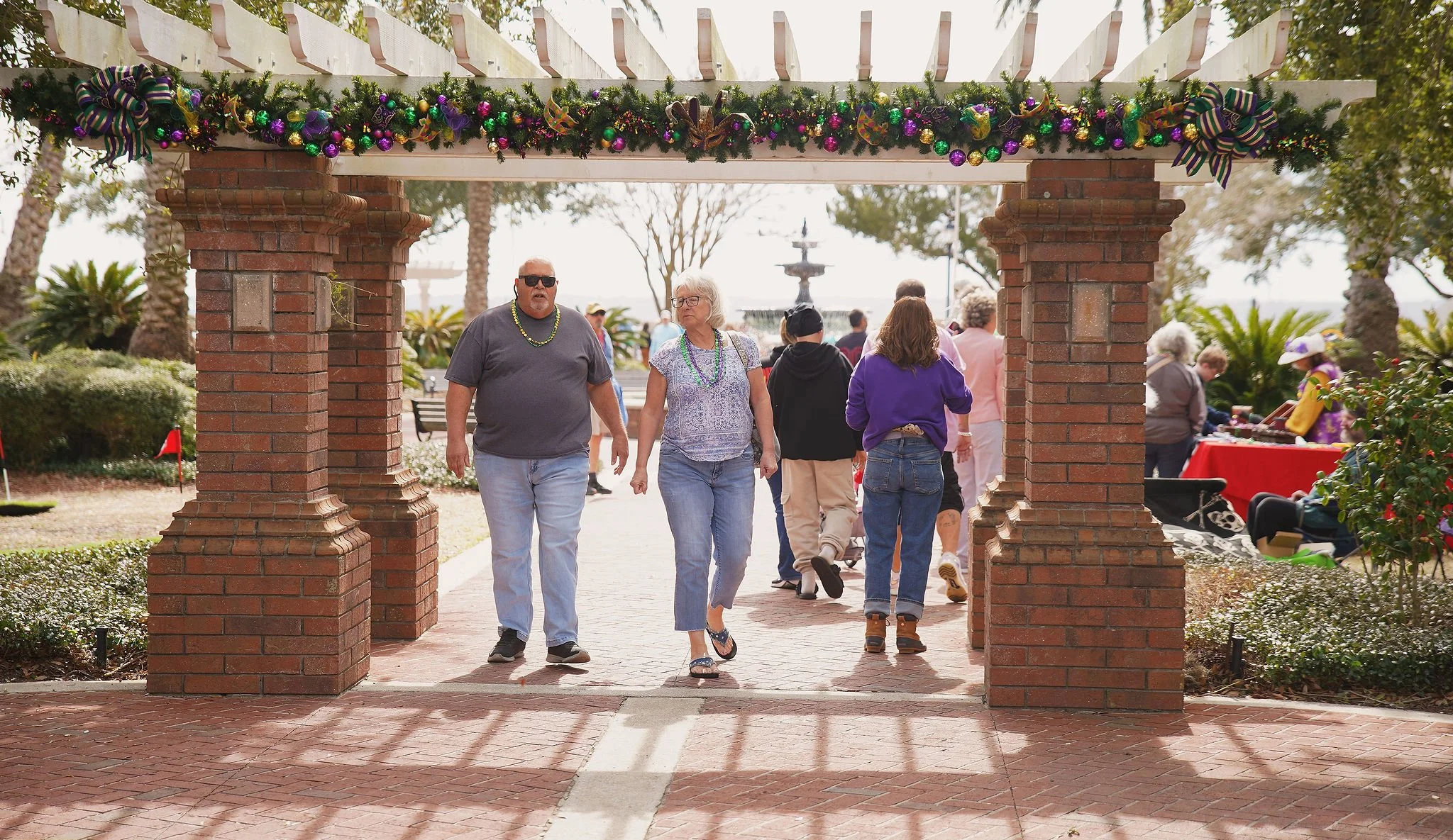 People walking under a decorated archway in an outdoor park with festive holiday decorations and a fountain in the background.