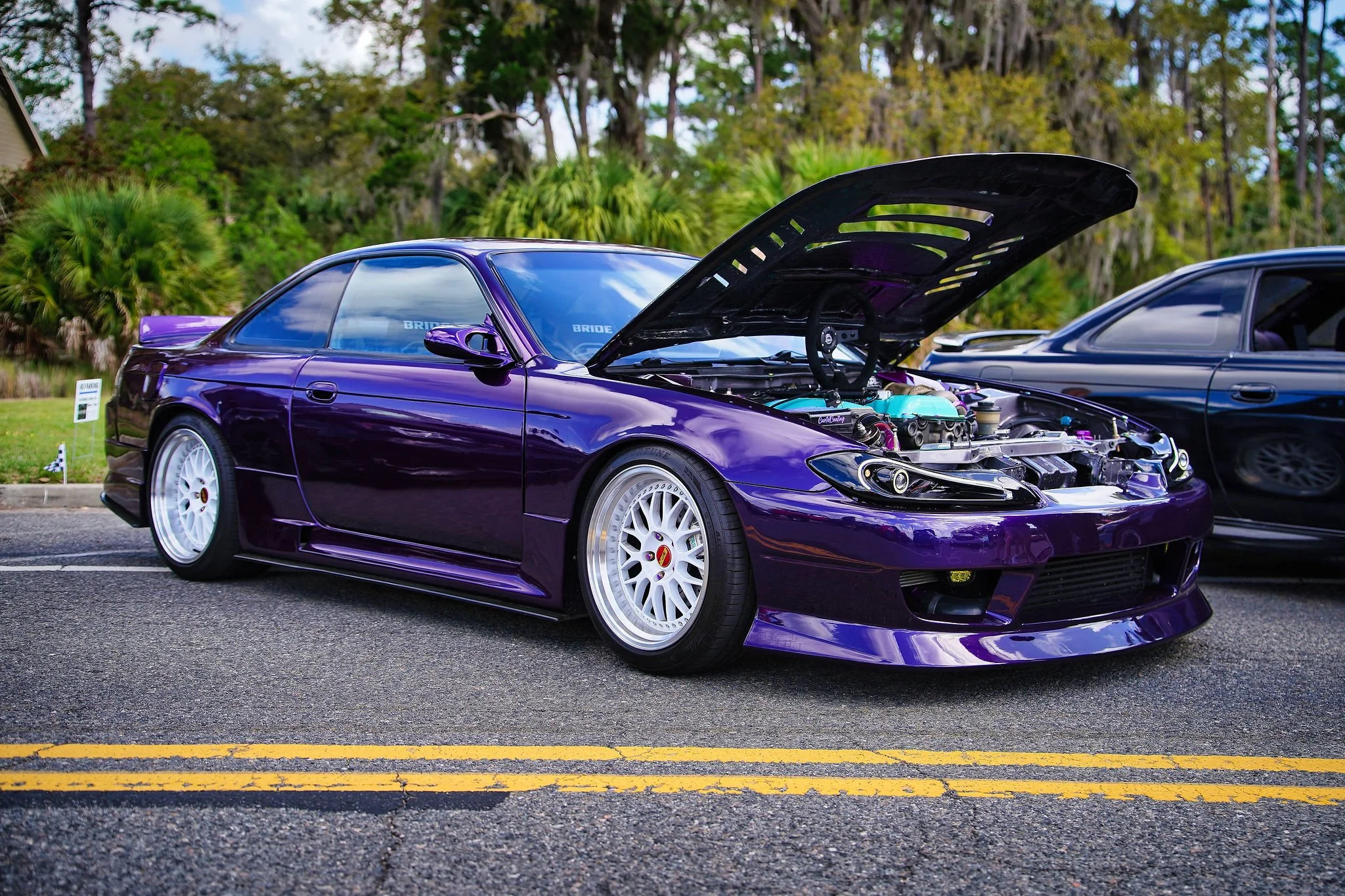 A purple modified sports car parked on the street with its hood open, showing the engine, next to another black car, with trees and a house in the background.