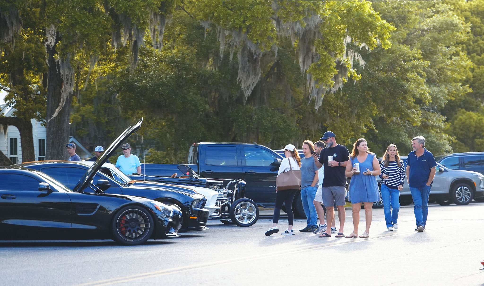 A group of people walking in a parking lot with cars, including a black sports car with its hood open, and trees with Spanish moss in the background.