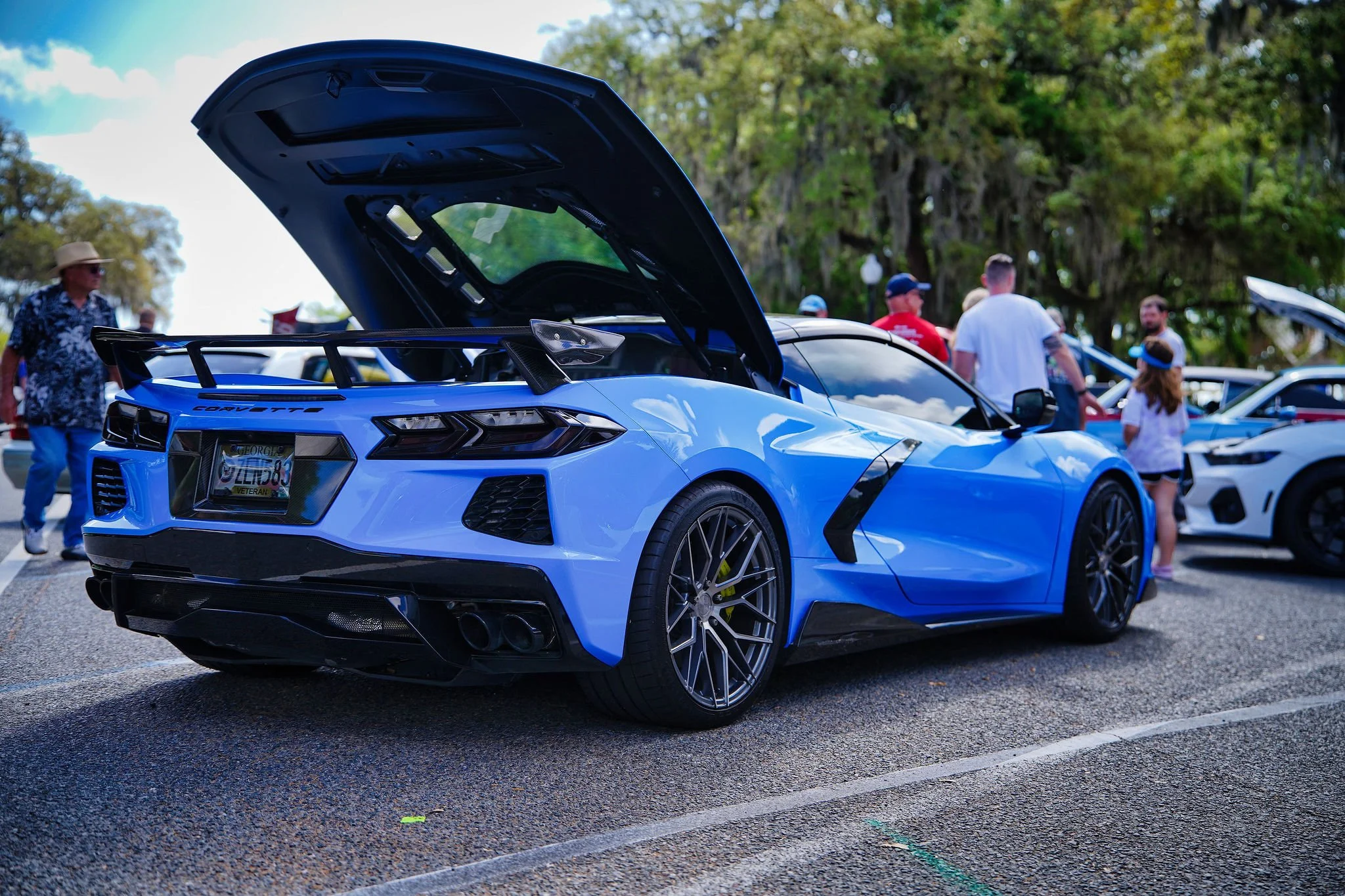 A blue Chevrolet Corvette with its hood open on display at a car show, with several people in the background.