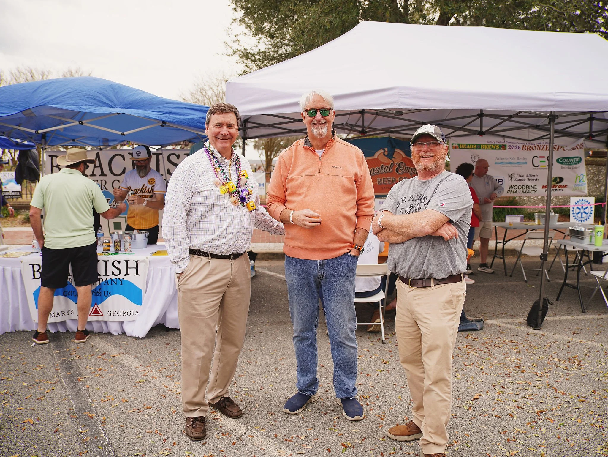 Three men standing together outdoors at a public event, smiling at the camera, with vendor tents and people in the background.