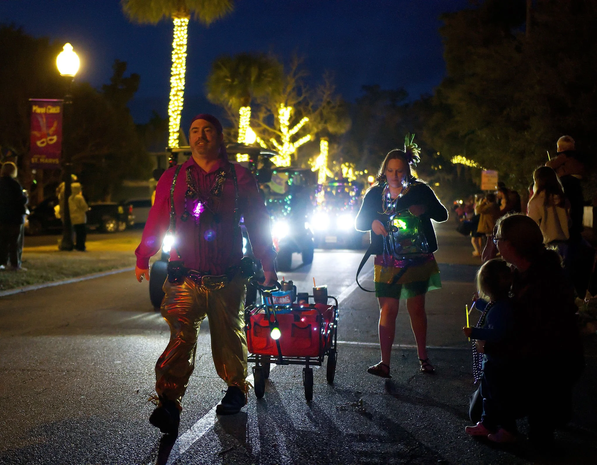 Nighttime street scene with people dressed in colorful, shiny costumes, some wearing accessories and beads, participating in a parade or festival. Bright lights and illuminated trees line the street.