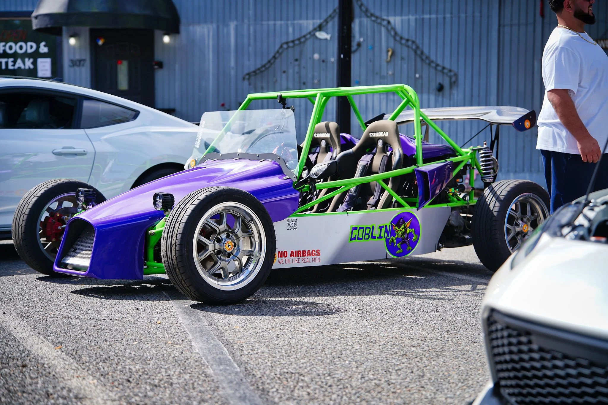 A purple and green open-wheel race car with racing seats labeled 'Corbeau' and a matchbox car logo that says 'Goblin', parked between two other cars in a parking lot.