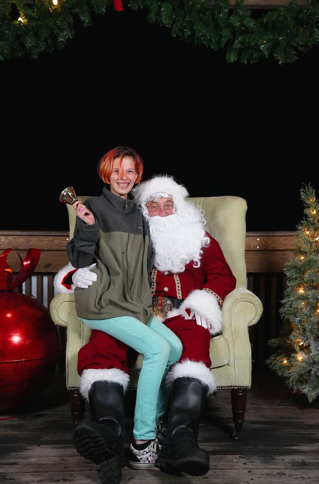A young girl sitting on Santa Claus's lap, holding a bell, and smiling in a Christmas setting with a decorated tree and gift bag nearby.