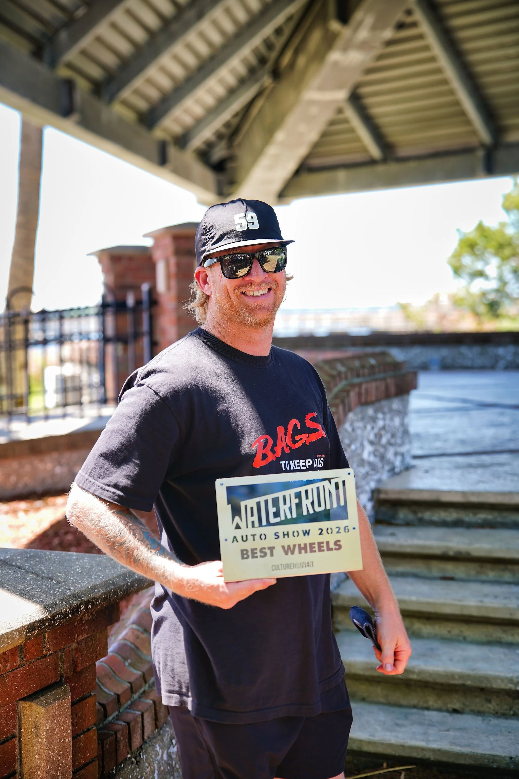 A man smiling and holding an award plaque that reads 'Waterfront Auto Show 2026 Best Wheels,' outdoors under a bridge, wearing a black cap, sunglasses, and a black T-shirt with red and white text.