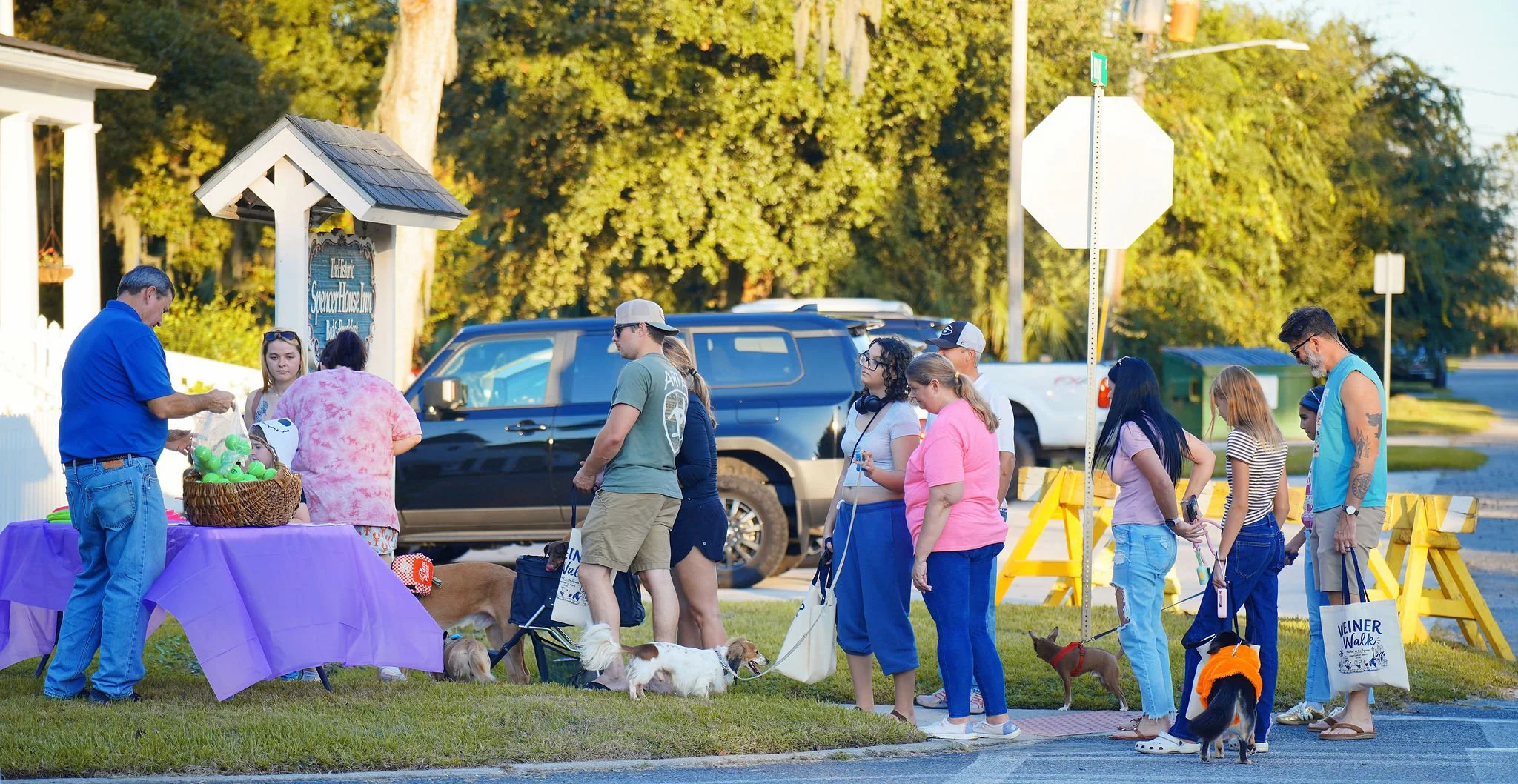 People standing in a line at an outdoor event with dogs, a vendor table, and a sign for Spencer House Inn in the background, with trees and parked cars around.