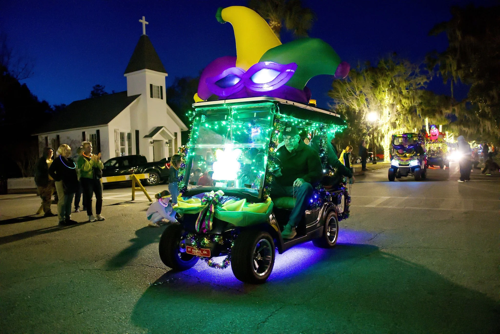 A decorated golf cart with Mardi Gras beads, lights, and a jester hat sculpture on top; parade spectators watch in the background during a nighttime parade in St. Marys, Georgia.