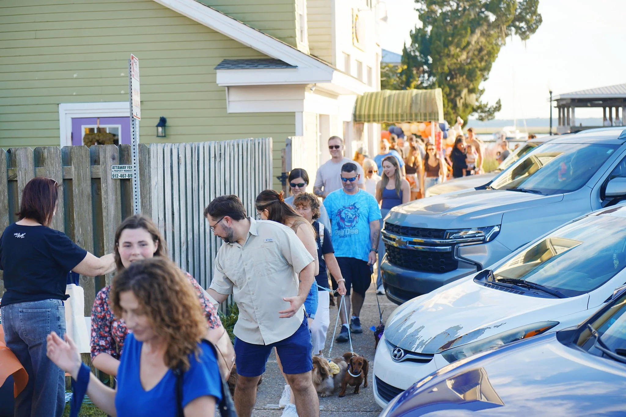 Crowd of people lining up outside a green building, with cars parked in front, during daytime.