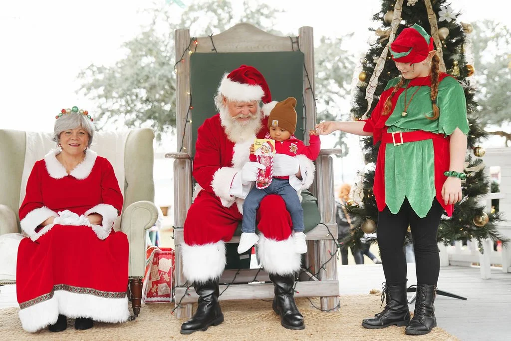 Santa Claus sitting on a throne holding a baby dressed in Christmas clothes, woman dressed as Mrs. Claus sitting nearby, and a young girl dressed as an elf standing next to a decorated Christmas tree.