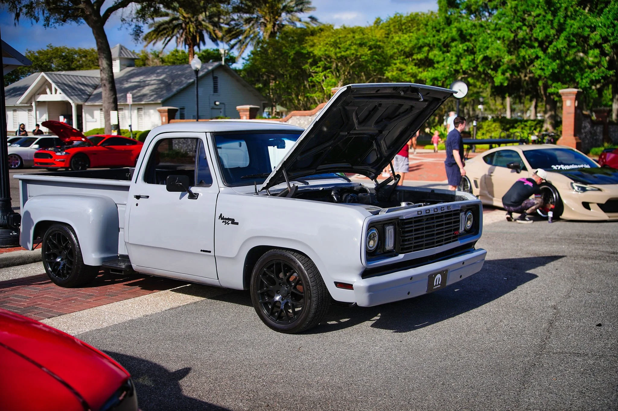 A white vintage Dodge pickup truck with its hood open, parked at a car show on a sunny day, with other cars and people in the background.