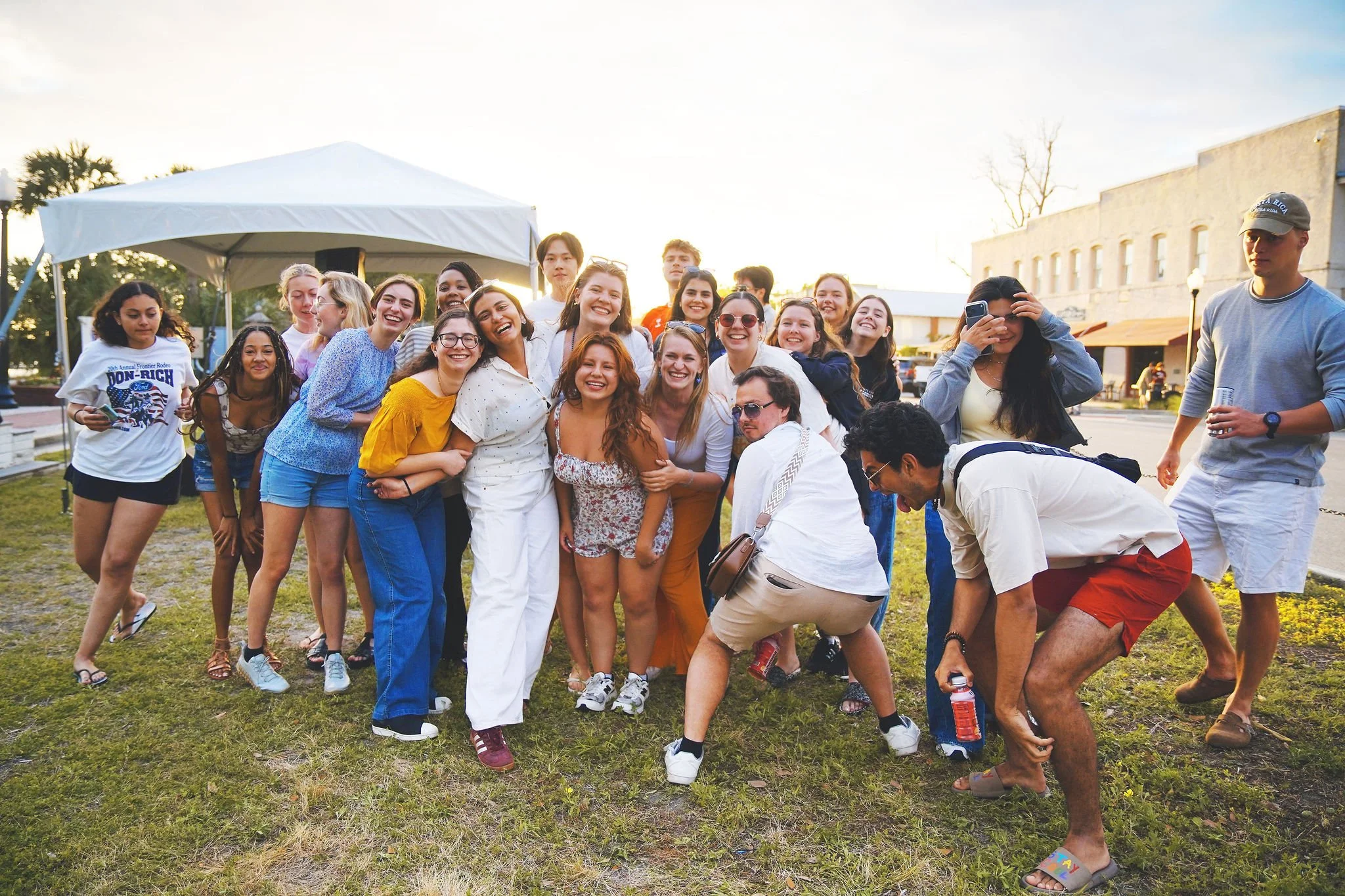 Group of young people smiling and posing outdoors on grass with a white tent and buildings in the background.