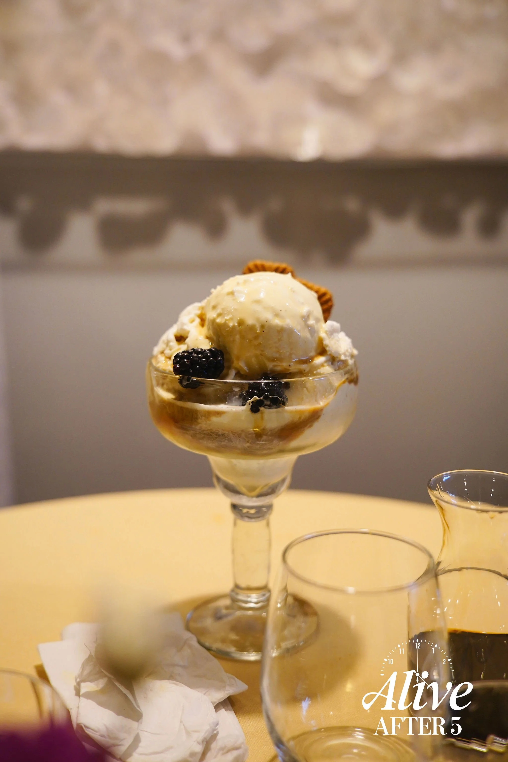 Dessert in a glass bowl with a scoop of vanilla ice cream, blackberries, and biscuits on top, placed on a table with empty drinking glasses and a creamer in the background.
