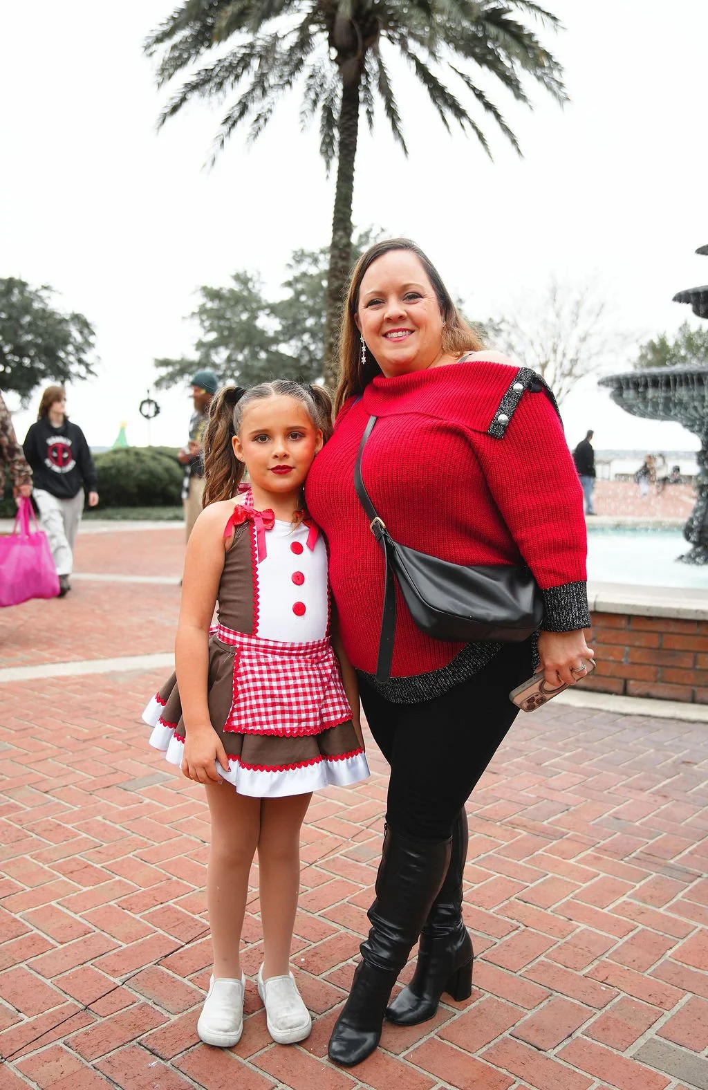 A woman and a young girl stand together outdoors, smiling for the camera near a fountain with a large palm tree behind them on a cloudy day.
