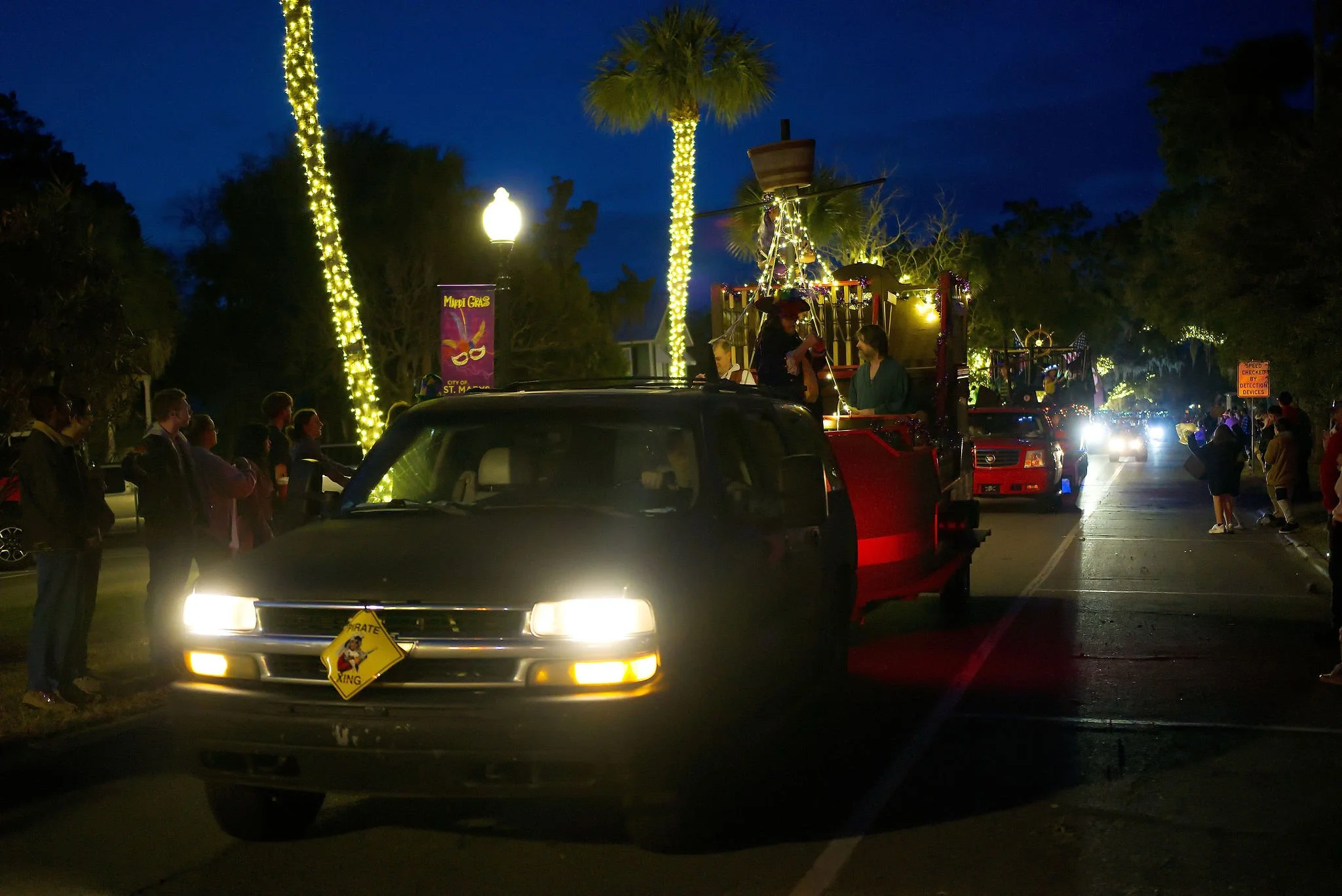 Nighttime parade with decorated float featuring people and palm trees lit with string lights, surrounded by spectators on street.