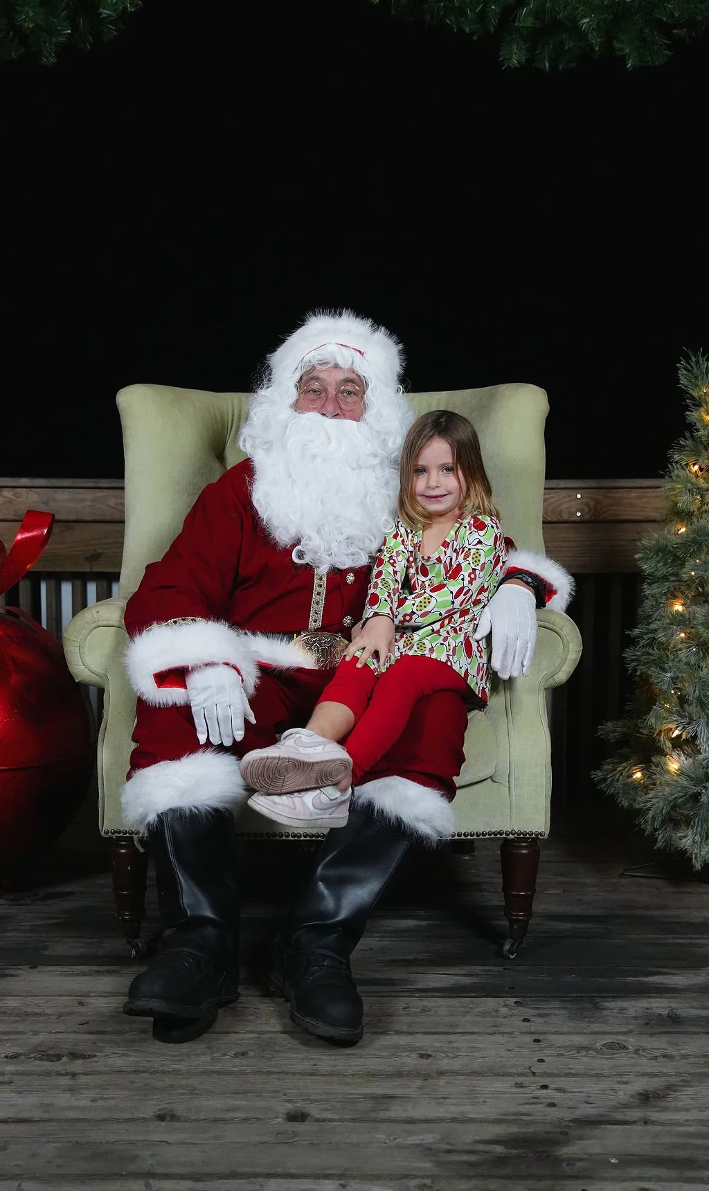 Santa Claus sitting in a green armchair with a young girl on his lap, both smiling, surrounded by Christmas decorations including a Christmas tree with lights on the right side of the image.