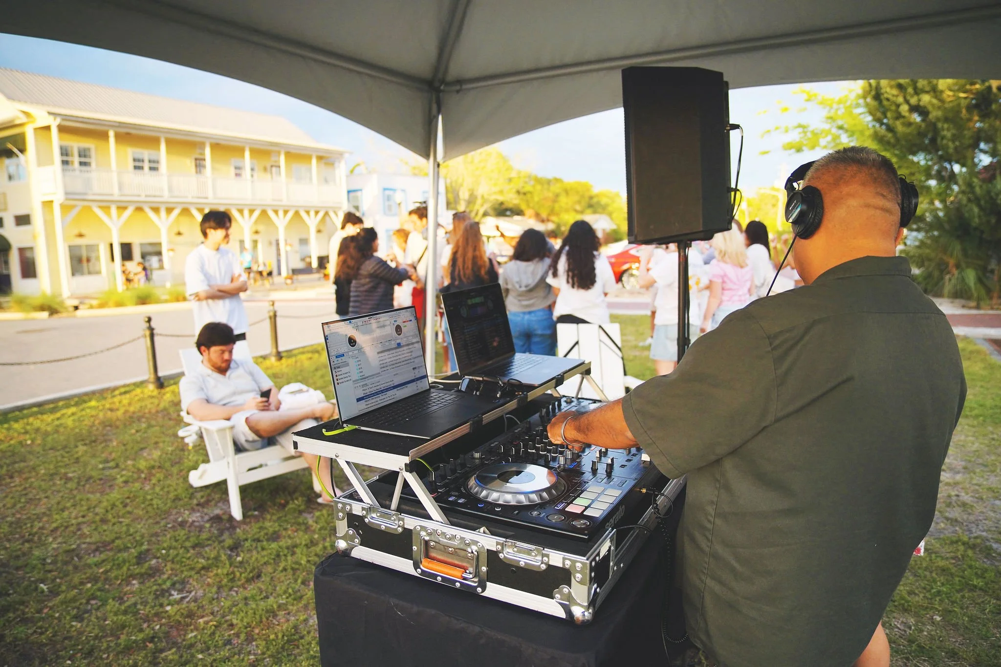 A DJ wearing headphones operates mixing equipment at an outdoor event, with a group of people gathered in the background under a tent.