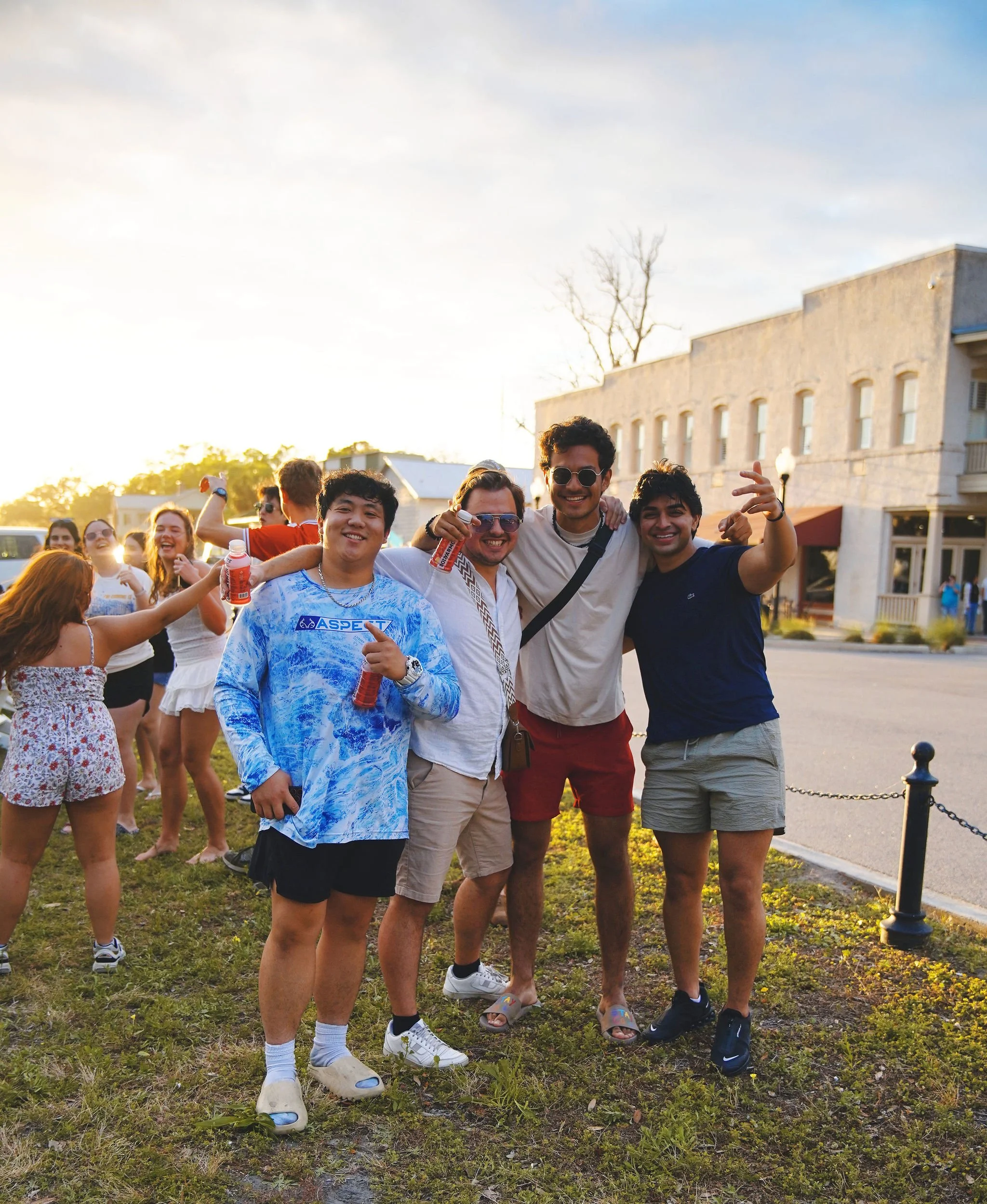 Group of young friends smiling, standing outdoors in front of a building during sunset, some holding drinks, celebrating and having fun.