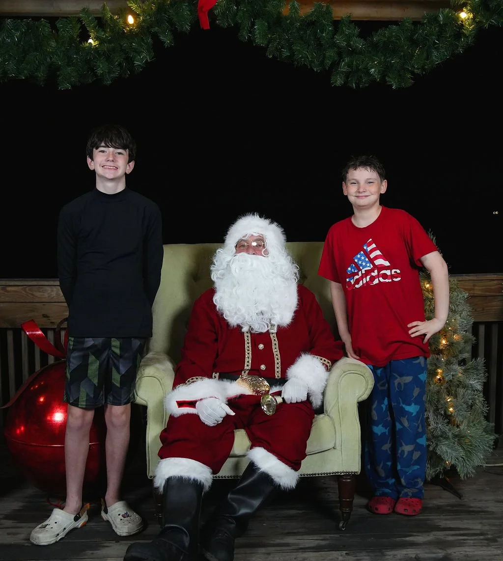 Two young boys standing next to Santa Claus who is seated on a green chair, with Christmas decorations including a large red ornament and a Christmas tree with lights and greenery in the background.