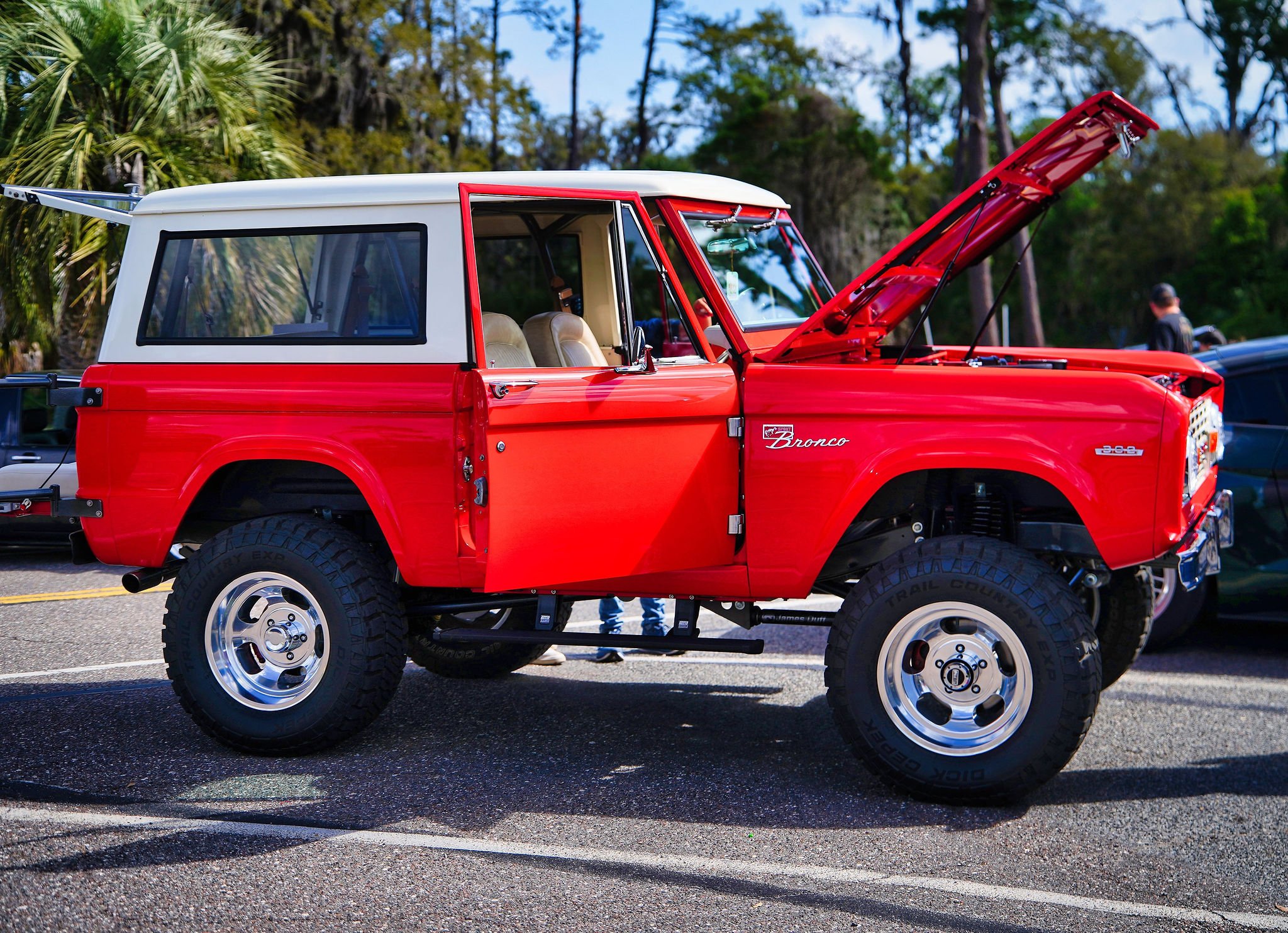 Vintage red Ford Bronco with its hood open, parked outdoors on a paved surface, with trees in the background.