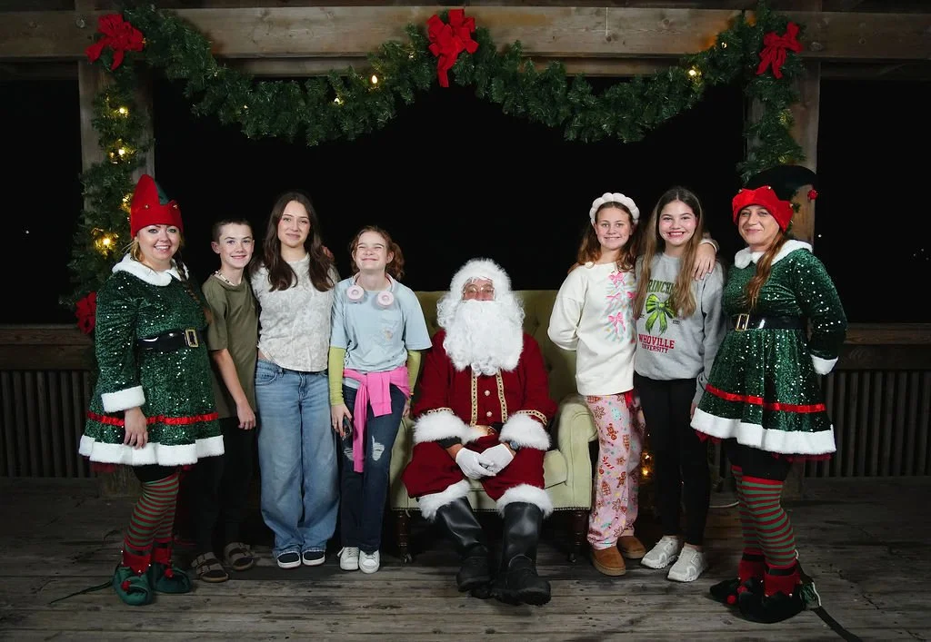 Group of children and two women dressed as elves with Santa Claus sitting in the center on a green chair, posing under a Christmas garland.