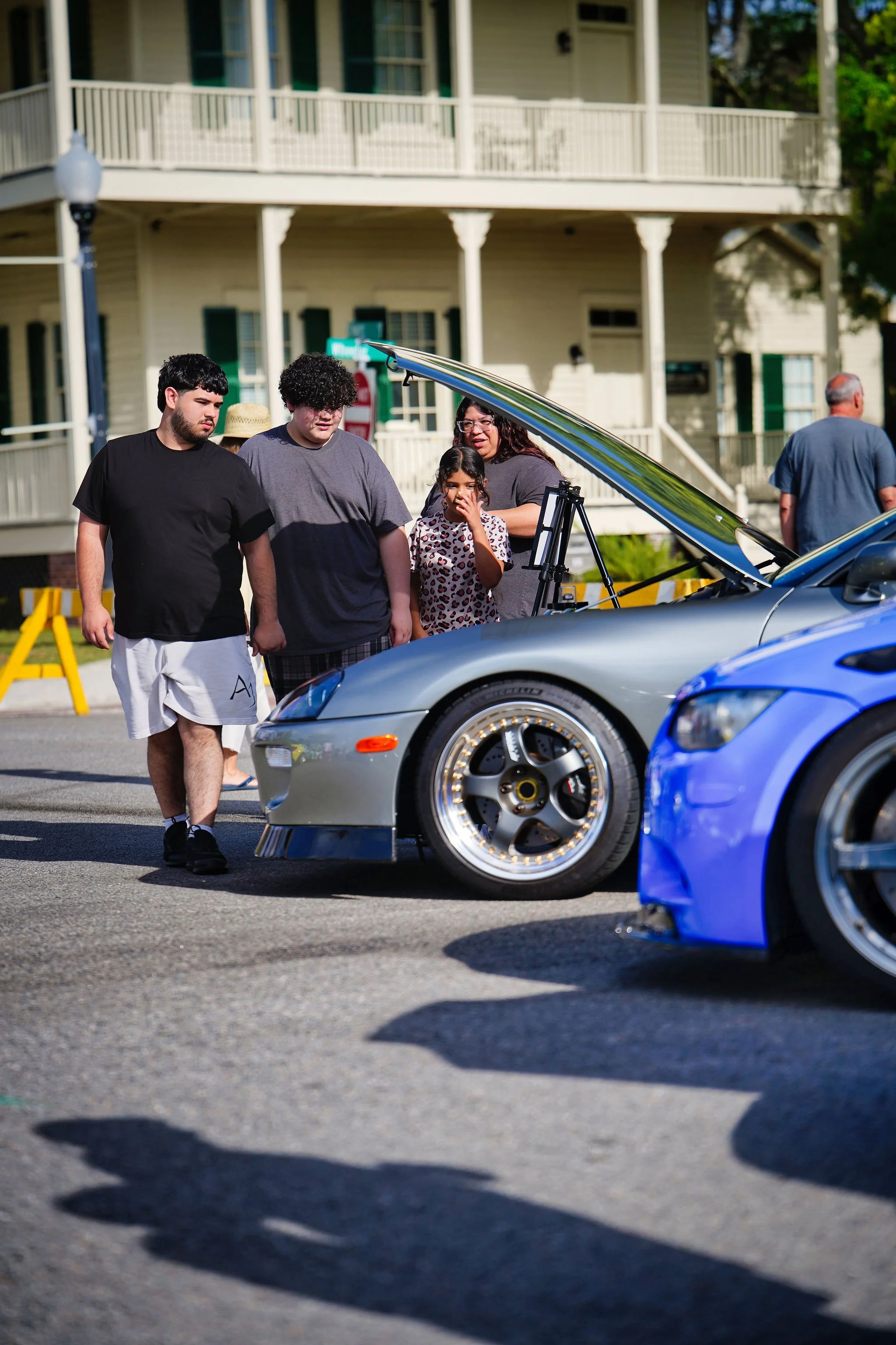 Group of people observing cars at an outdoor car show, with a house in the background.