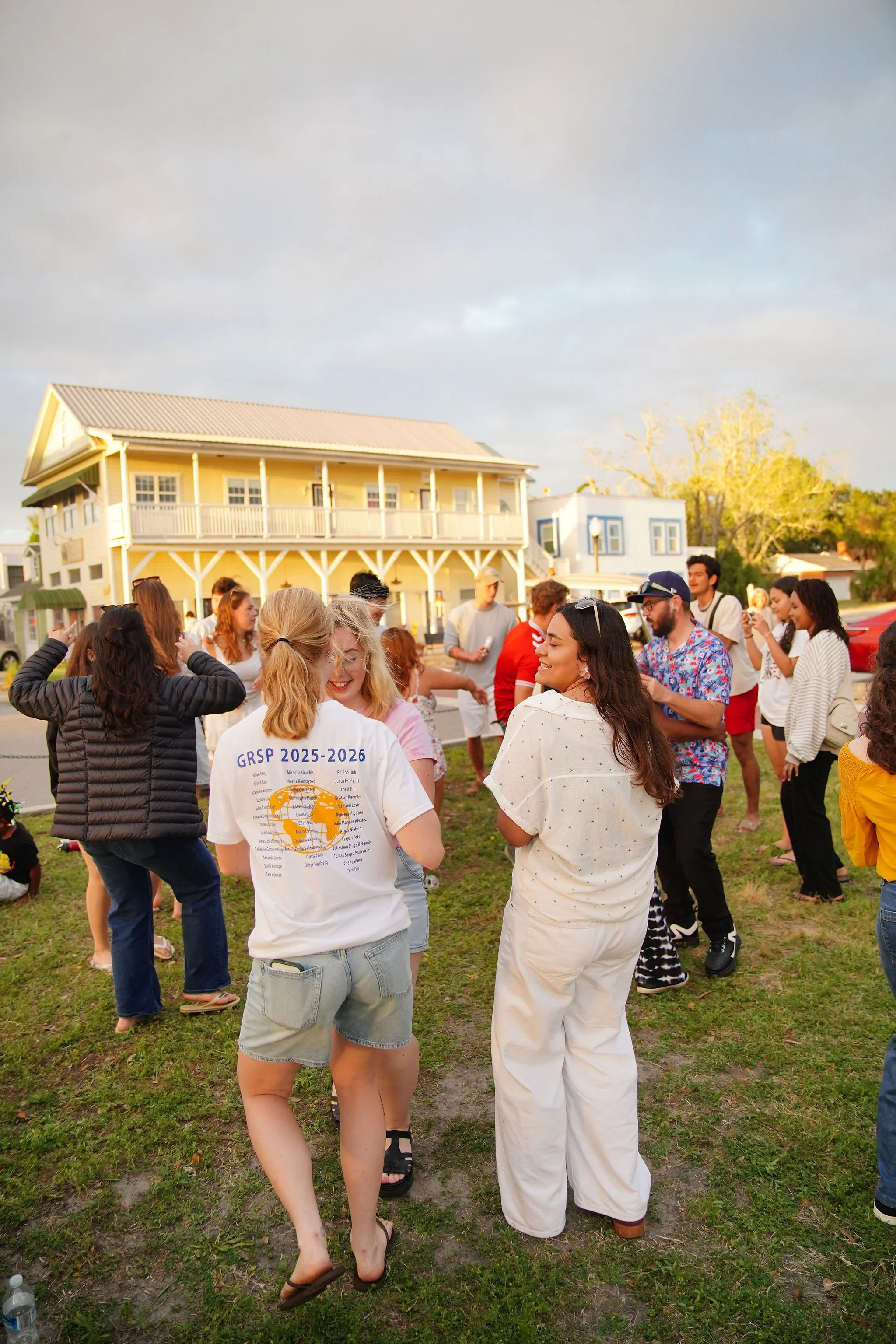 A group of people gathered outdoors on a grassy area in front of a yellow building with a balcony, enjoying a social event during the daytime.