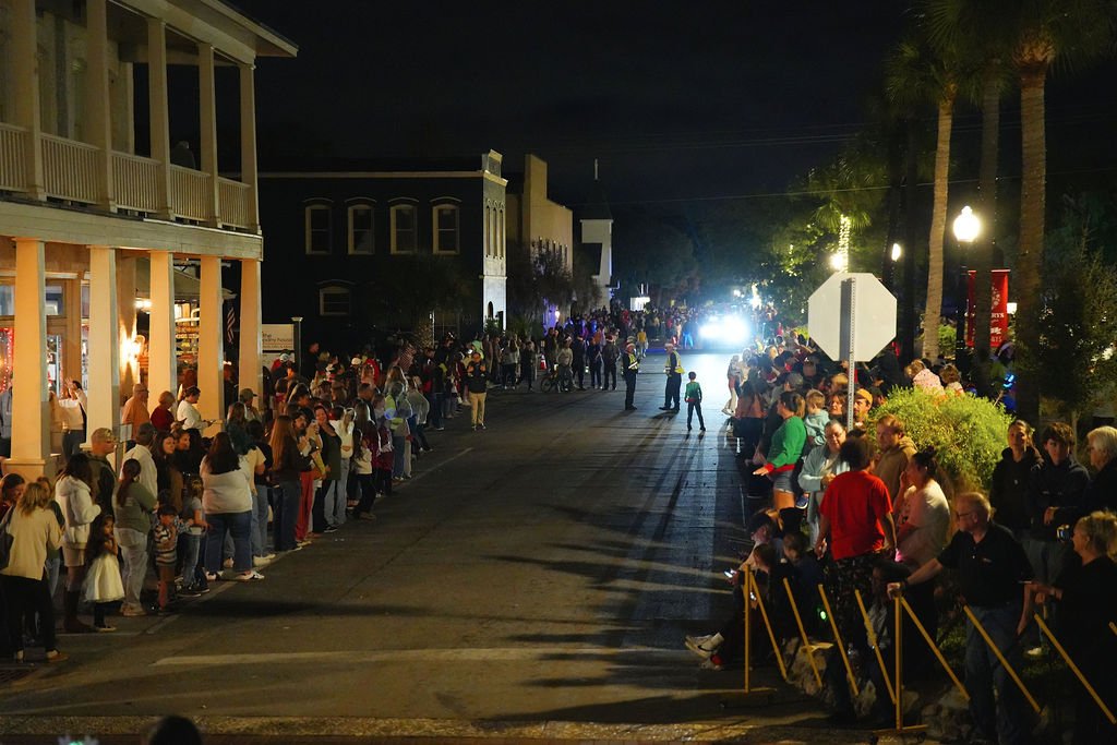 People lined up along both sides of a street at night, waiting for a parade or event, with some children and families visible and bright lights in the background.