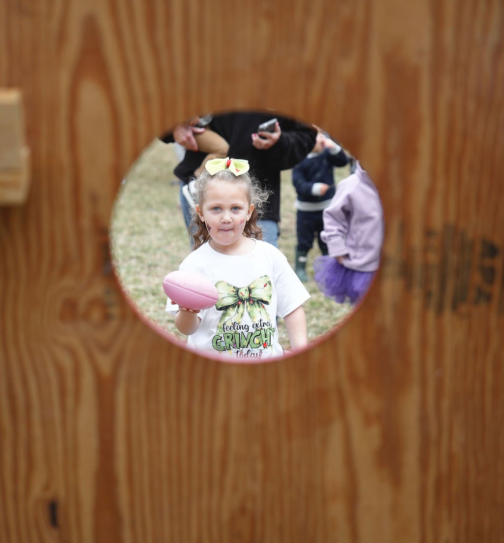 Girl with a bow in her hair holding a pink egg, viewed through a circular hole in a wooden surface, with other children in the background.