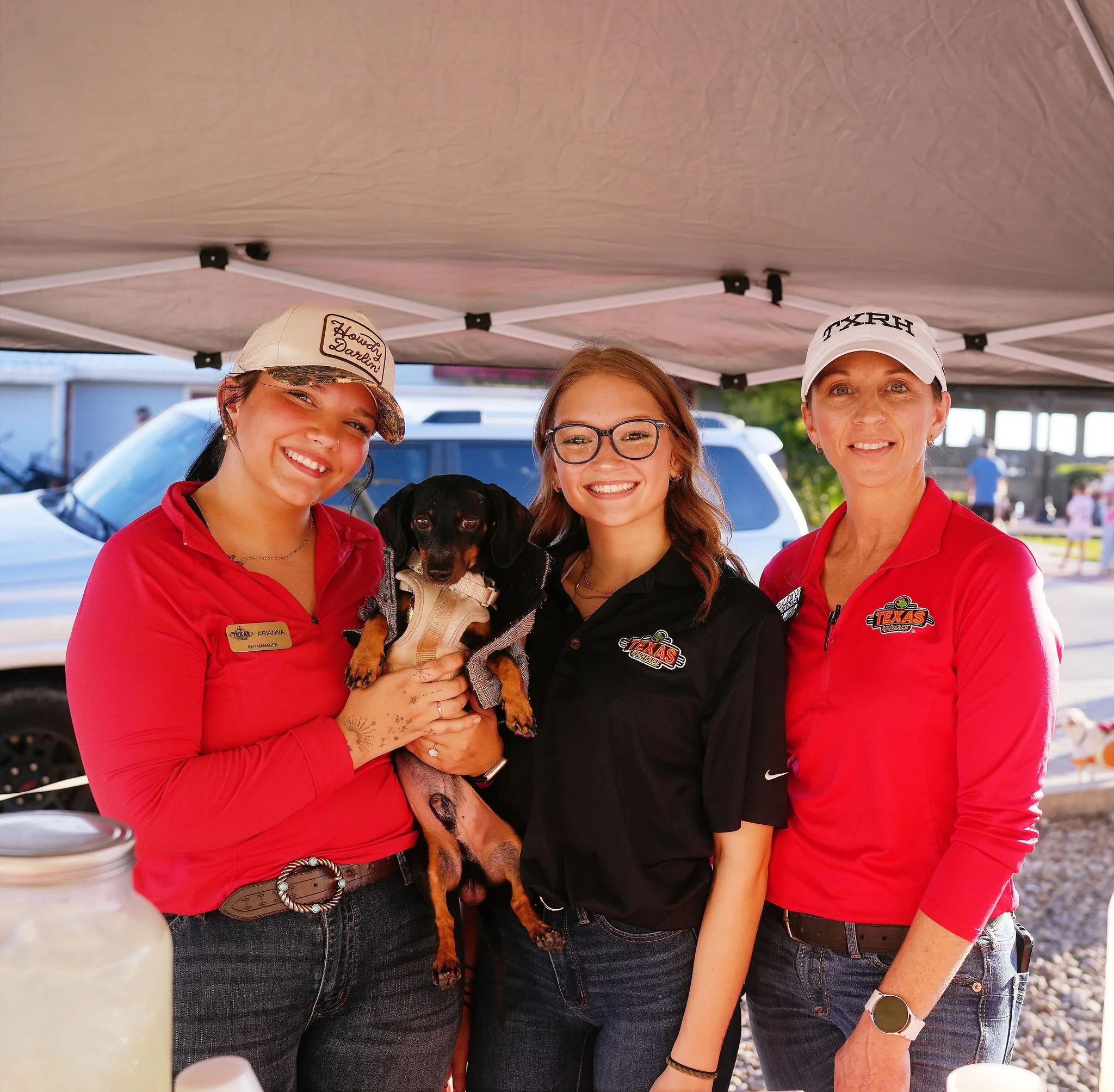 Four women standing under a canopy, smiling, with a dog held by one of the women. The women are wearing red and black shirts with logos, and one woman has a badge that reads 'Arianna Key Manager'. The women appear to be at an outdoor event, with a pa