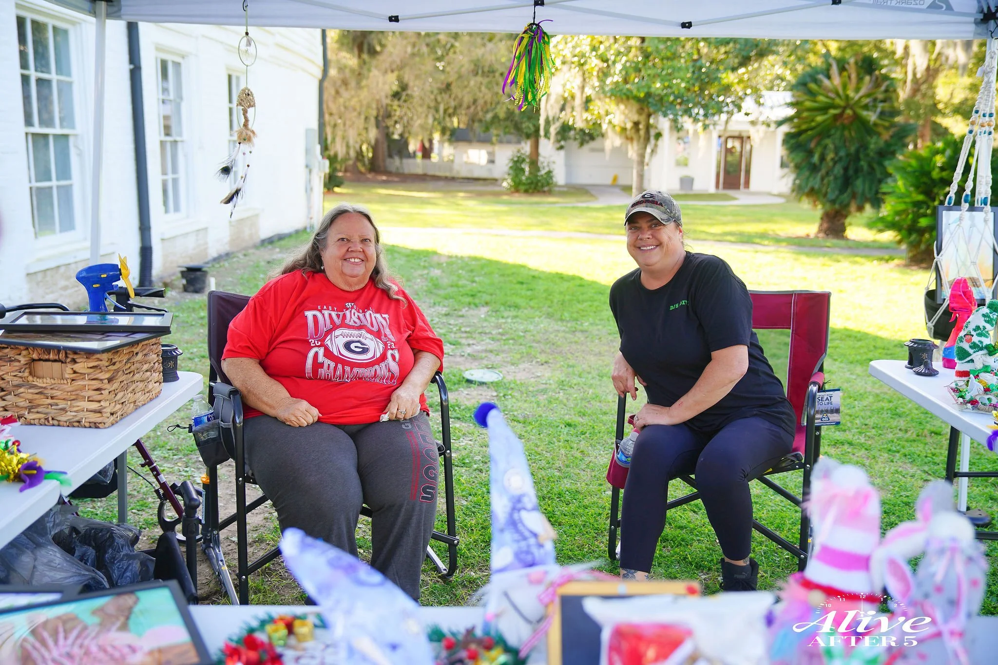 Two women sitting outdoors under a canopy, smiling at each other with tables of craft items and decorations around them, in a grassy yard with trees and houses in the background.