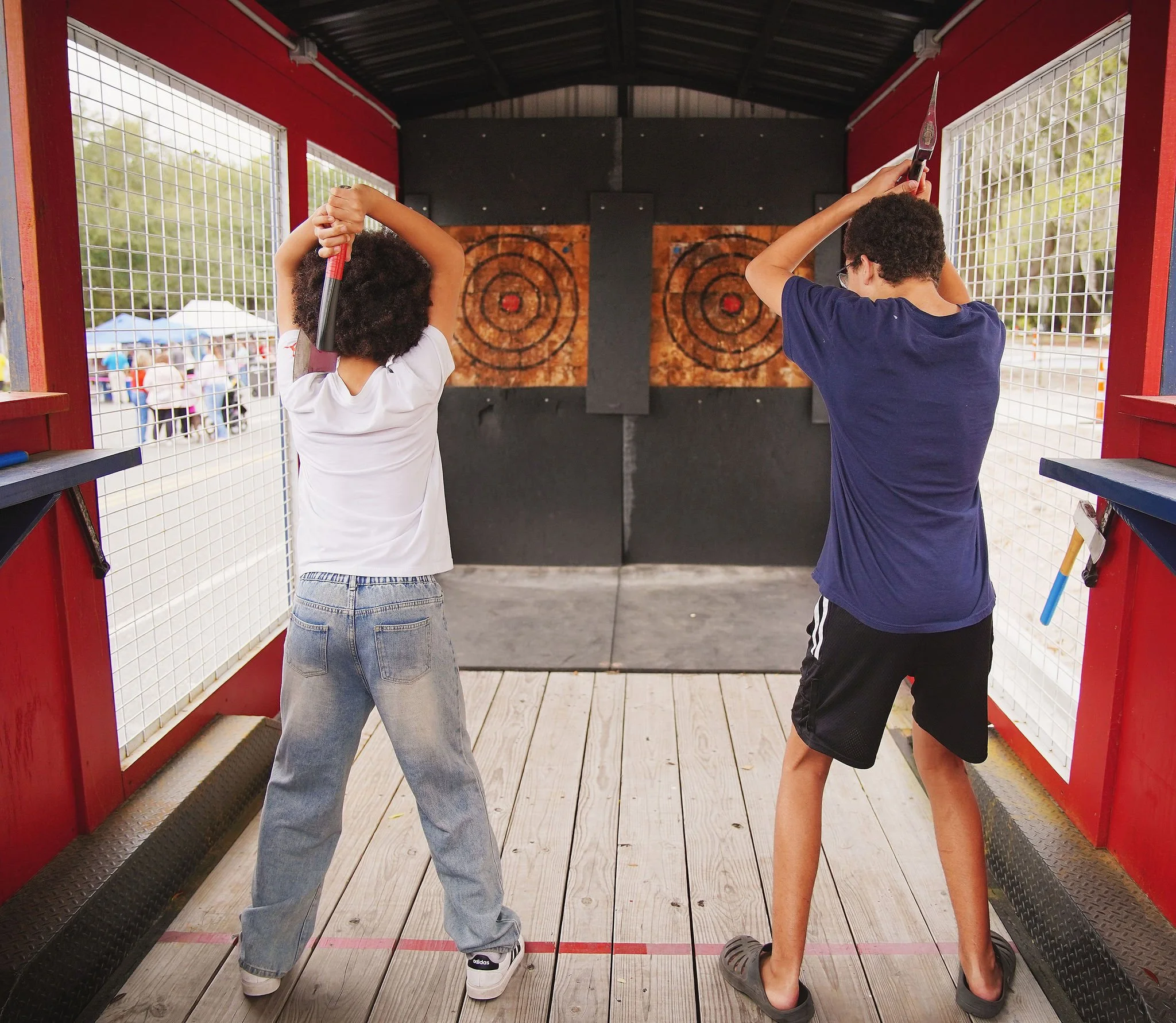 Two kids practicing axe throwing at an outdoor target range.