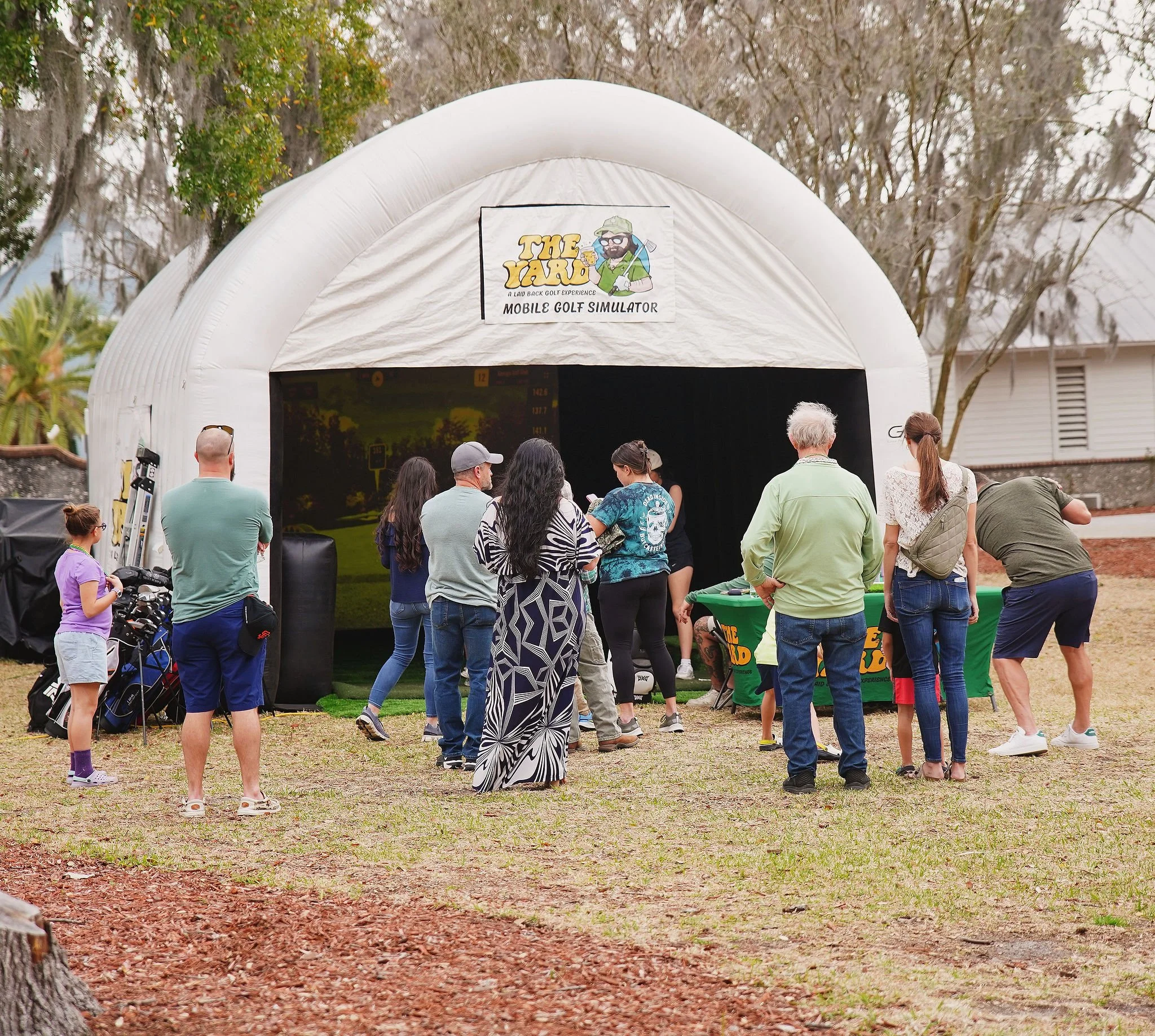 People gathered outside a white tent with a sign that reads 'The Yard', which is a mobile golf simulator.