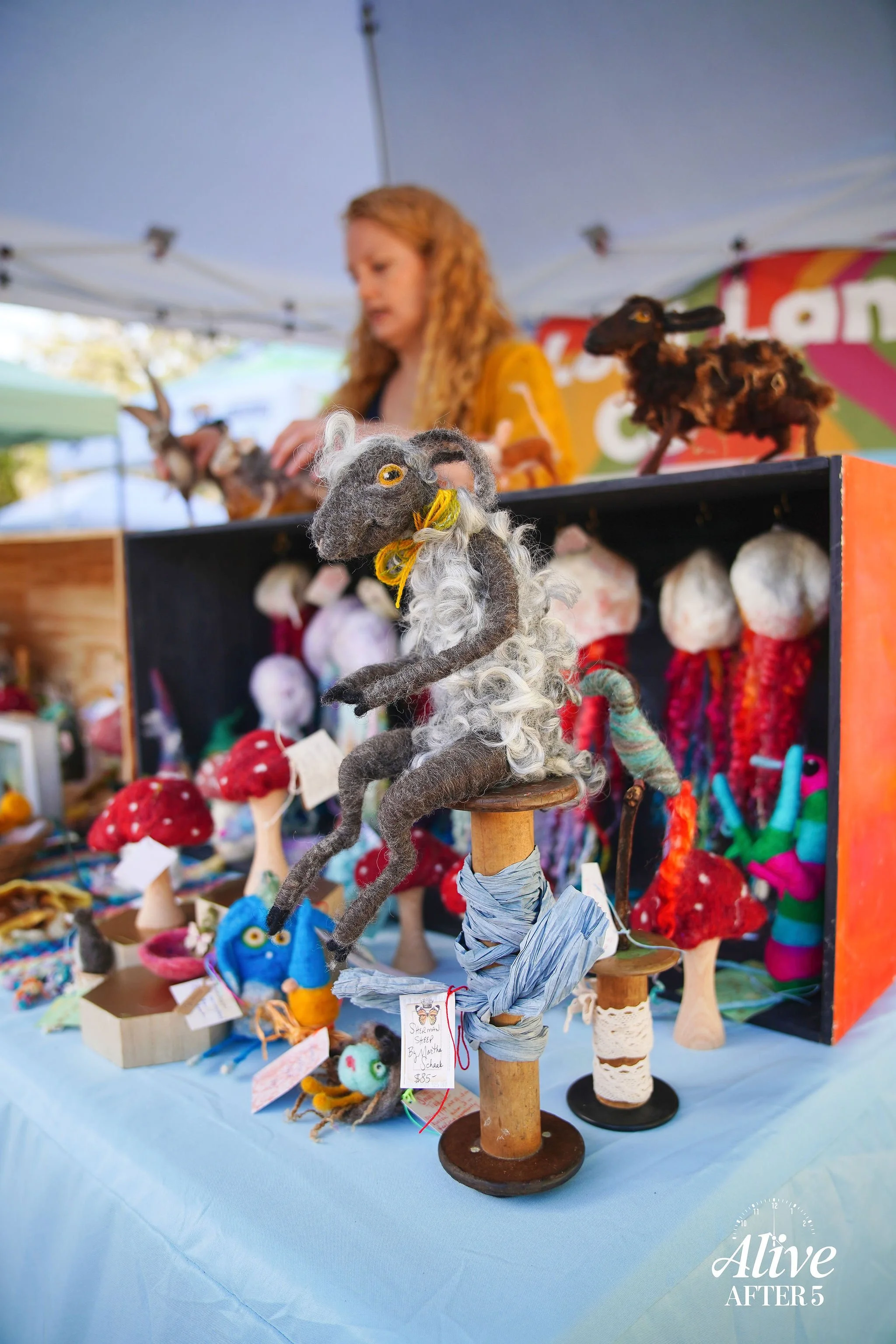 A felted animal sculpture resembling a horse with a mane, sitting on a wooden stand at a craft fair stall with other felted animals and handmade crafts displayed on the table behind it.