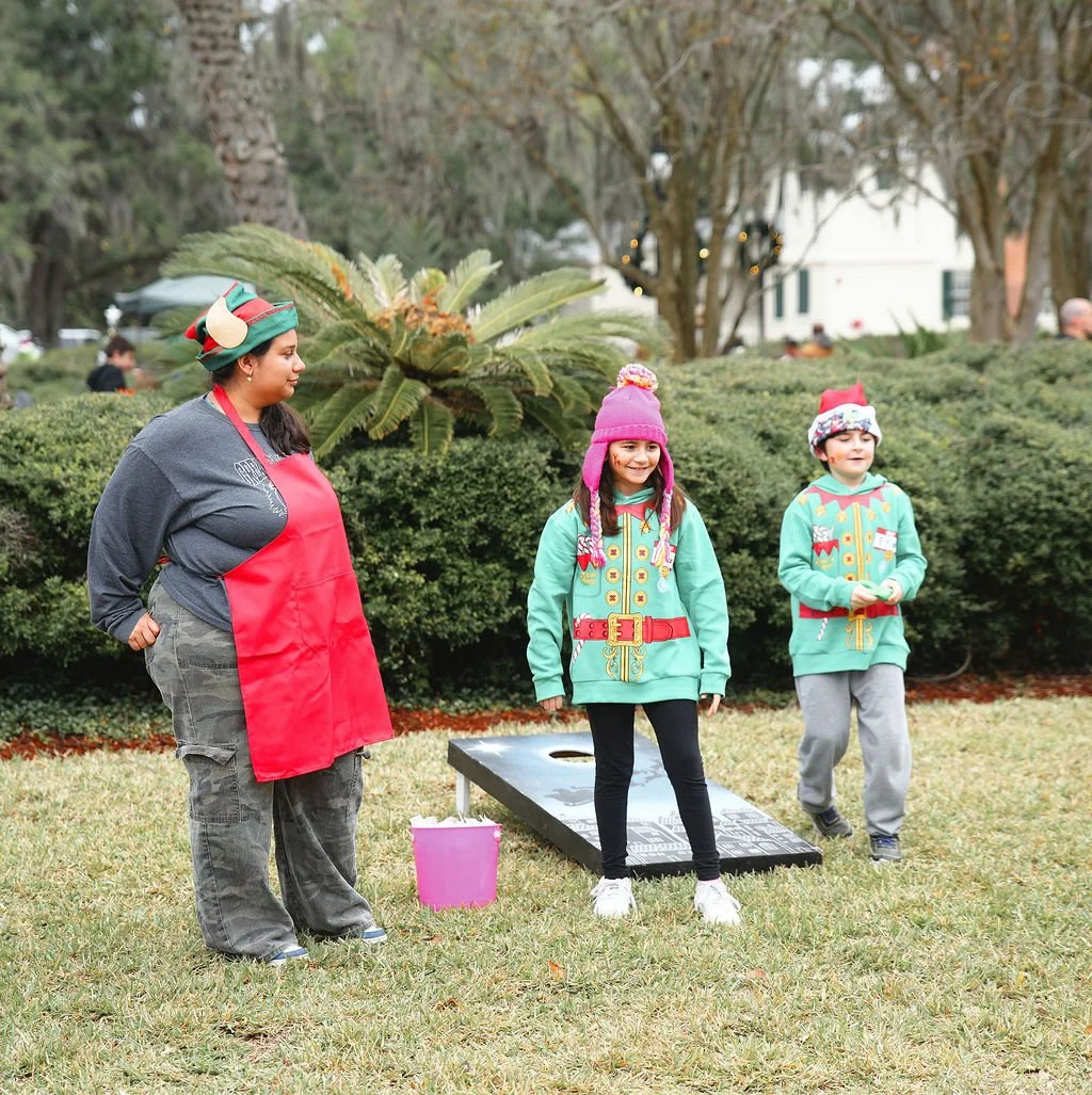 Two children dressed in Christmas-themed sweaters and hats playing a game of cornhole outdoors, with a woman watching beside them, in a park or garden setting during daytime.