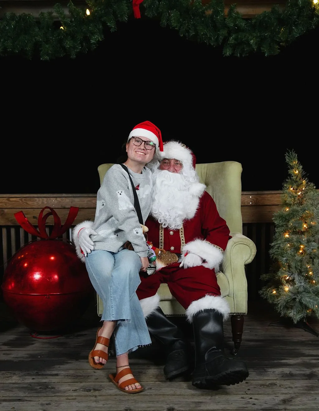 A young woman sitting on Santa Claus's lap at a Christmas event, both wearing festive hats and smiling. Santa is dressed in a red suit with white trim, sunglasses, and black boots, seated in a green armchair. There is a decorated Christmas tree on th
