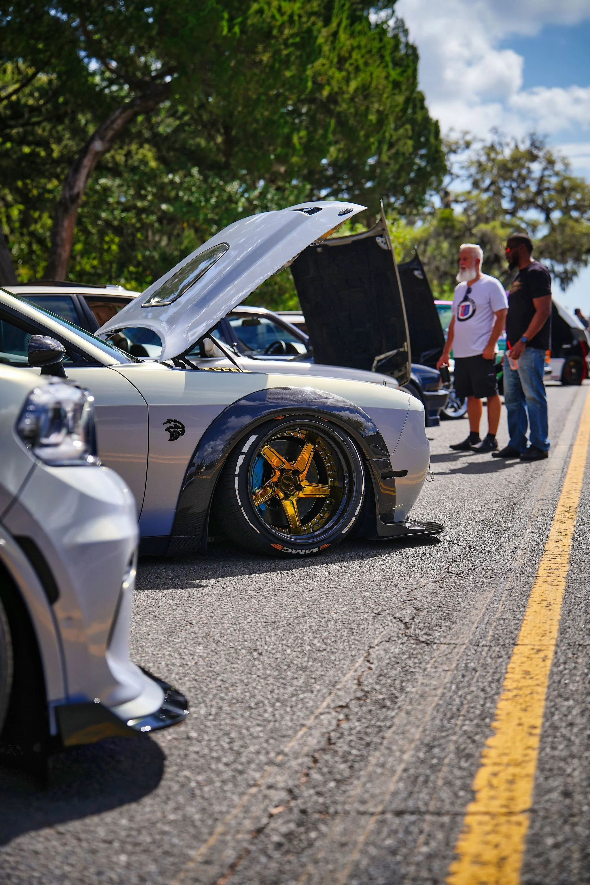 A close-up of a silver sports car with custom gold wheels parked on the street at an outdoor car show with people walking and cars in the background.