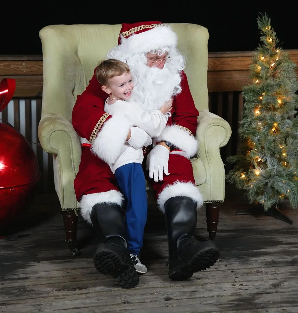 A young boy hugging Santa Claus sitting on a green armchair, with a decorated Christmas tree to their right and large red Christmas ornament to their left.