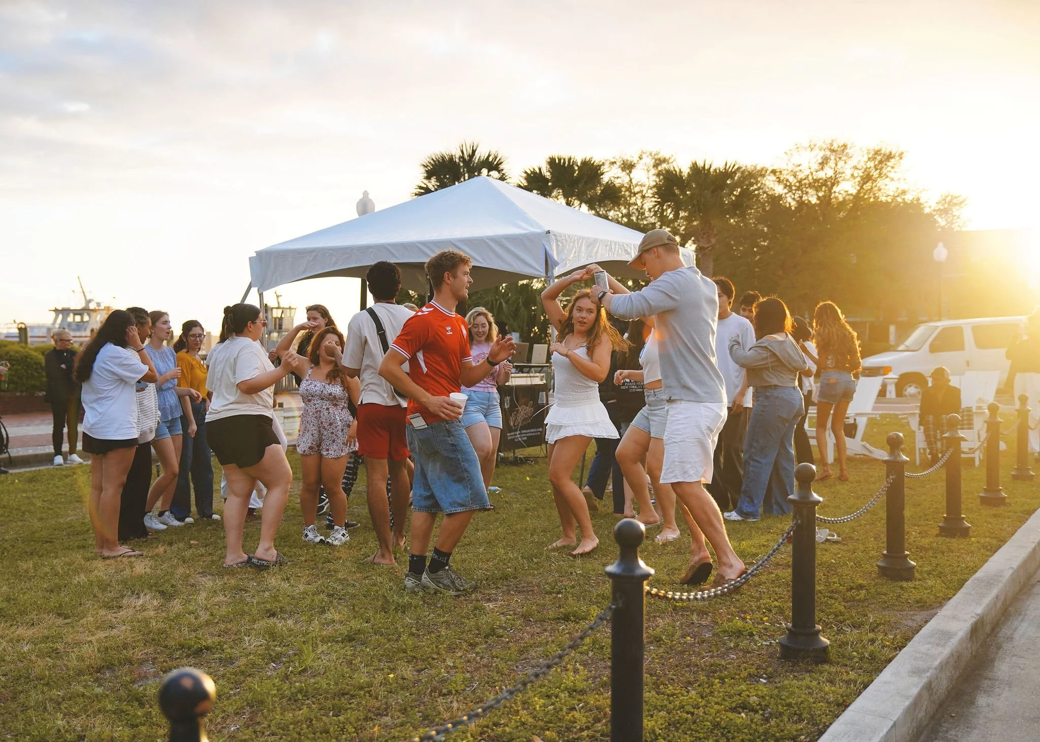 People dancing and socializing outdoors at sunset near a white tent, with palm trees, boats, and a fence in the background.