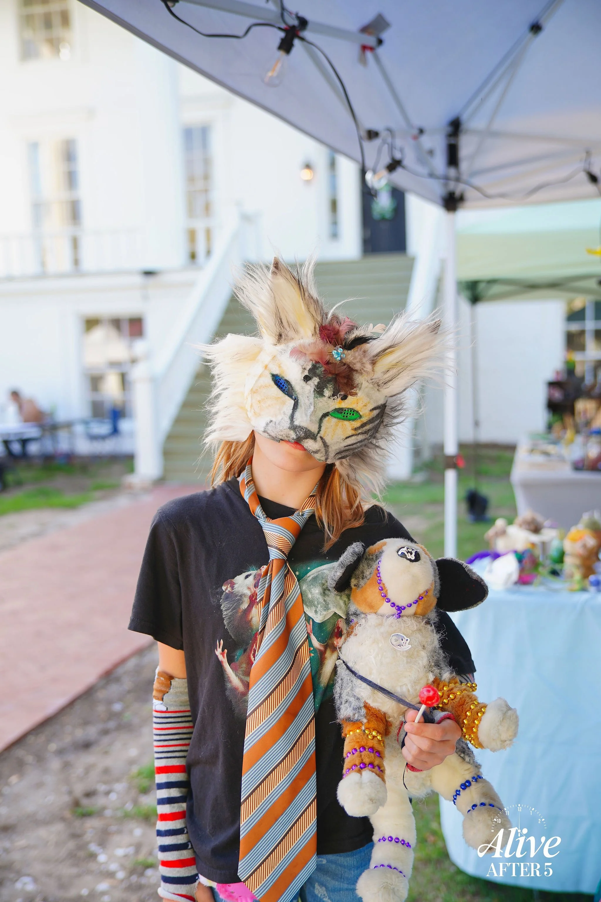 Person wearing a cat mask and holding a stuffed dog toy at an outdoor event with vendor tents in the background.