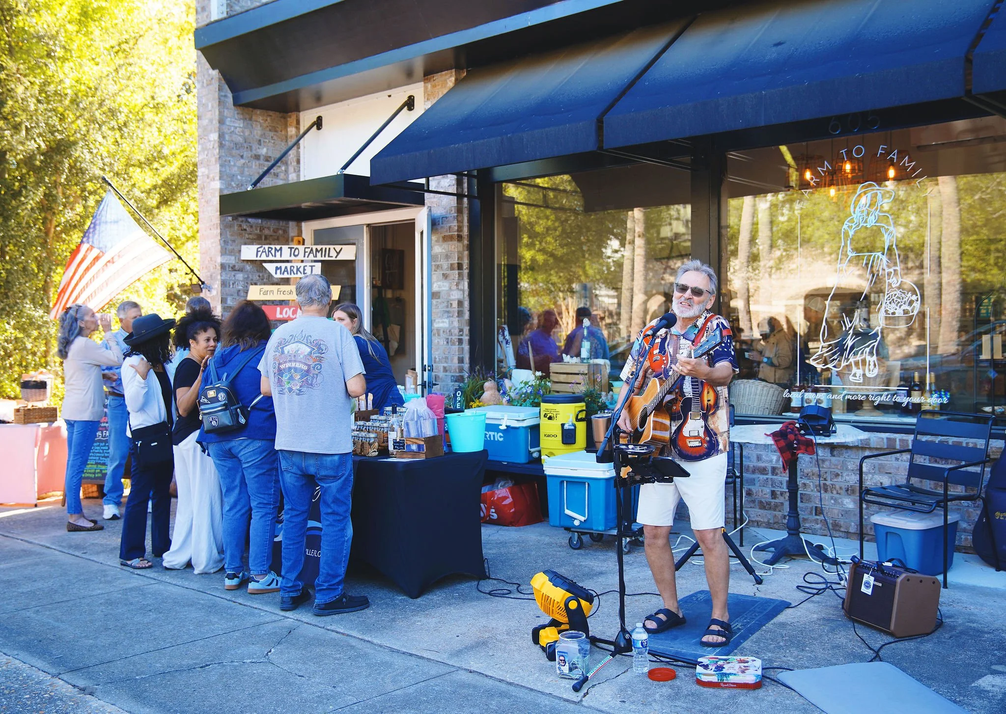 A street market scene with people waiting in line at a vendor stall labeled 'Farm to Family Market'. A man is performing with a guitar, singing into a microphone, wearing sunglasses, a colorful shirt, and white shorts, with musical equipment around h