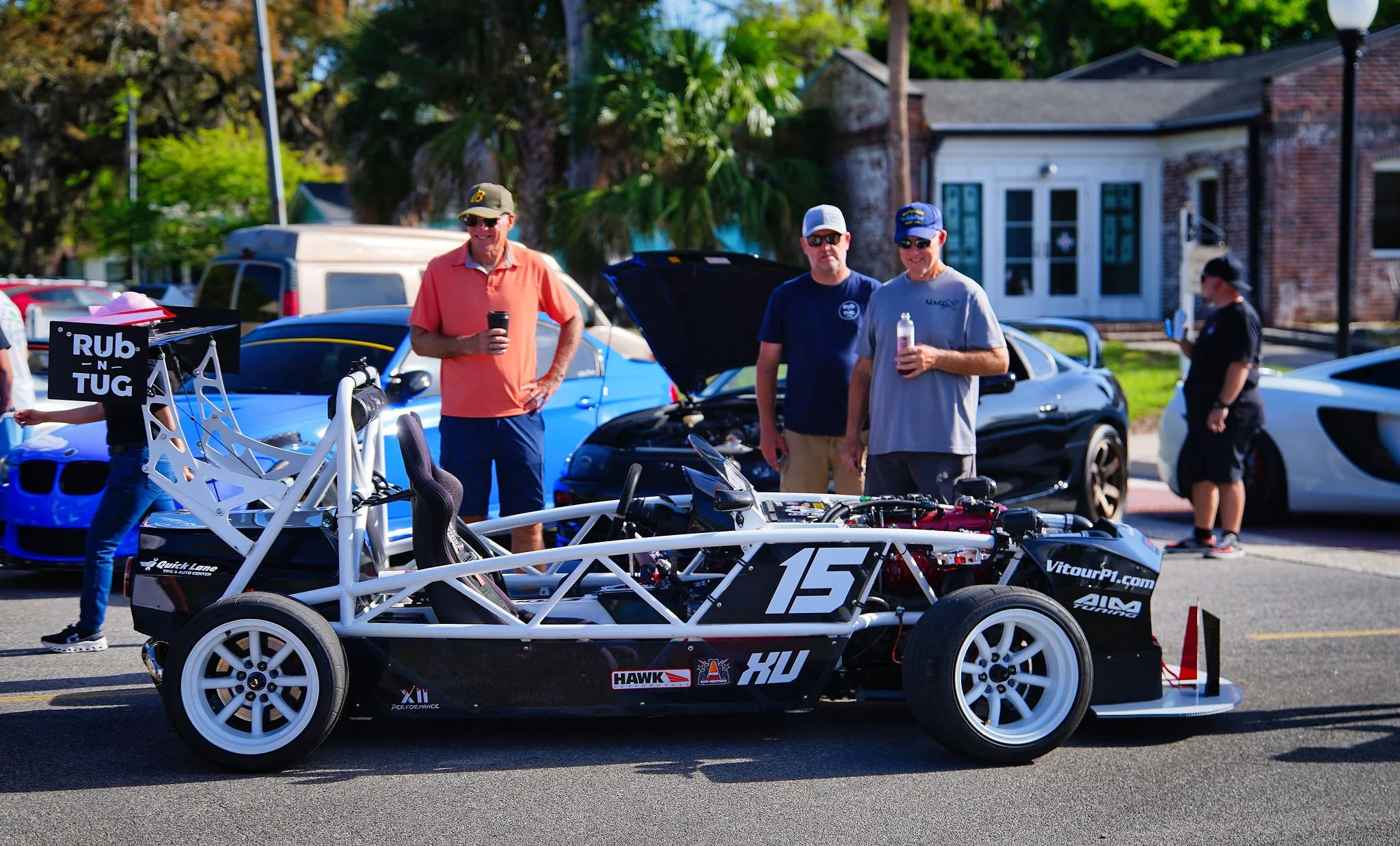 A race car with open framework parked on the street, surrounded by people, some holding drinks, with cars in the background and trees.