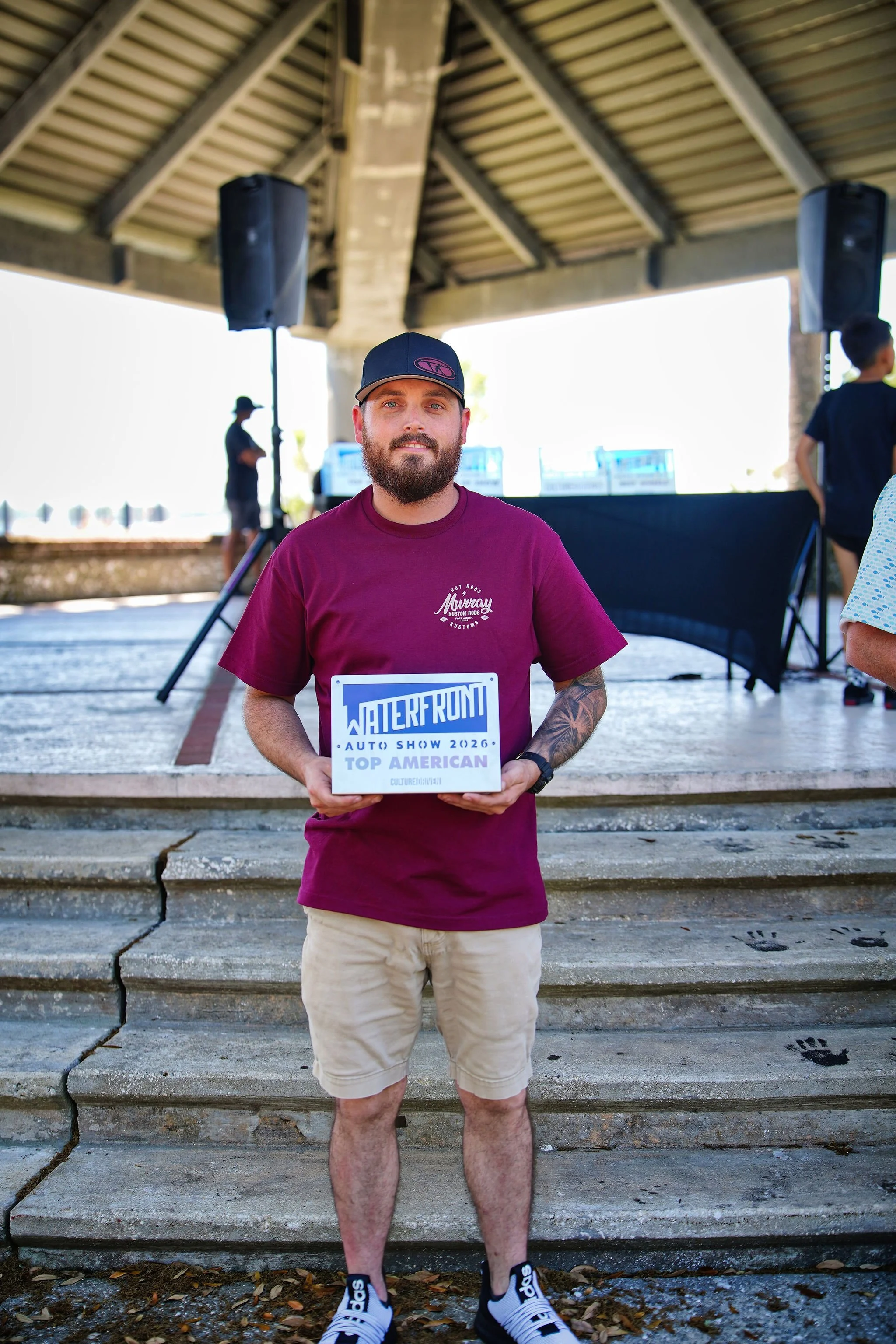 A man with a beard and tattoos is holding a sign that says, 'INTERFRONT AUTO SHOW 2026 TOP AMERICAN,' standing outdoors on a stone staircase under a pavilion. In the background, there are speakers, a DJ setup, and other people.