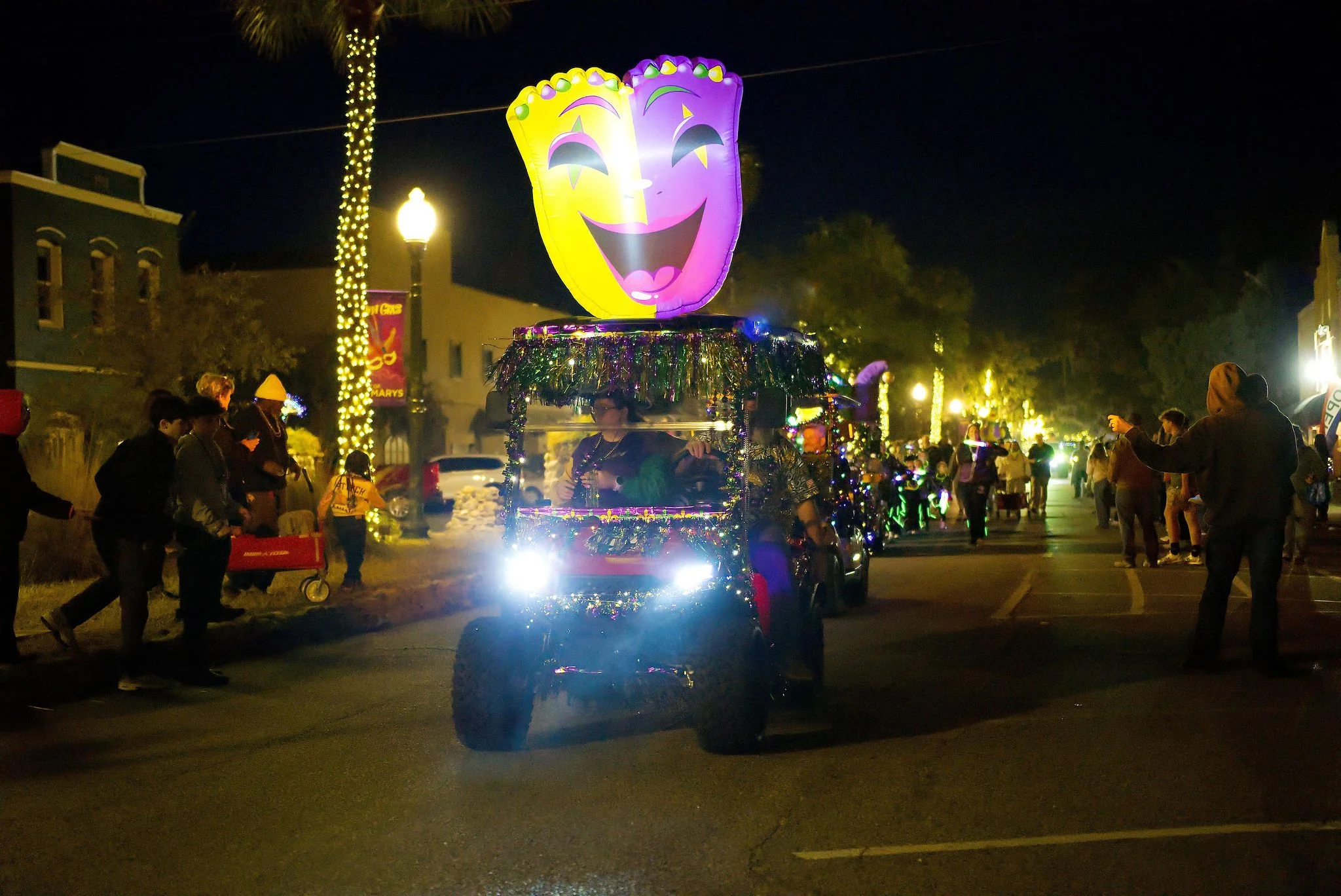 Nighttime parade with decorated golf carts featuring large cheerful face balloons on top, surrounded by people and string lights on trees.