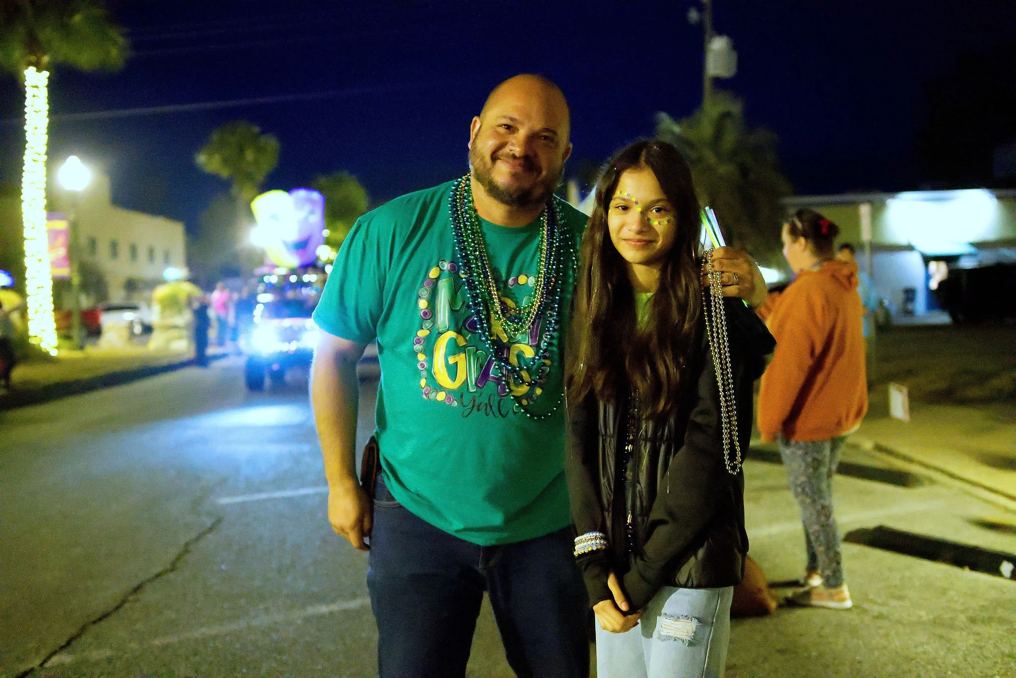Two people, a man and a woman, smiling and posing together at night outdoors with carnival or festive lights in the background. The man is wearing a green shirt with beads around his neck, and the woman is wearing a black jacket and light jeans. Ther