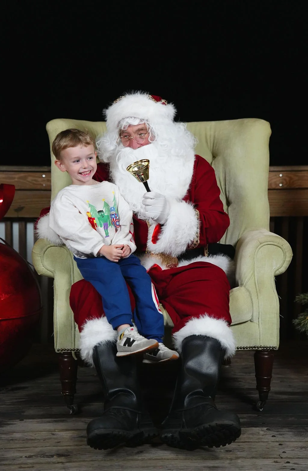 A young boy sitting on Santa Claus's lap, smiling and wearing a white sweater with colorful Christmas characters, in a festive setting.