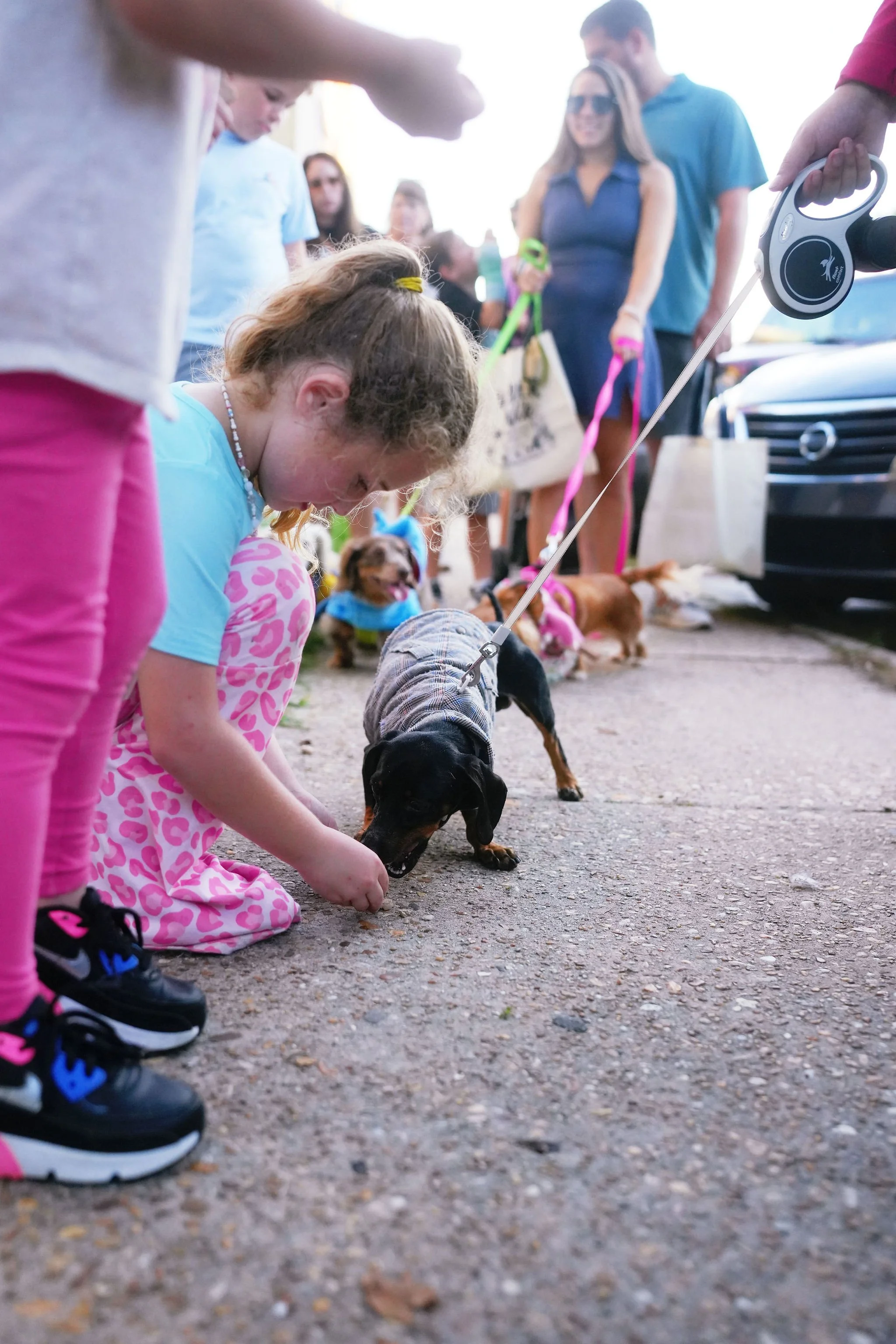 Children and adults with dogs at a pet-friendly outdoor gathering, with people standing and socializing, some holding leashes and bags, near a black car.